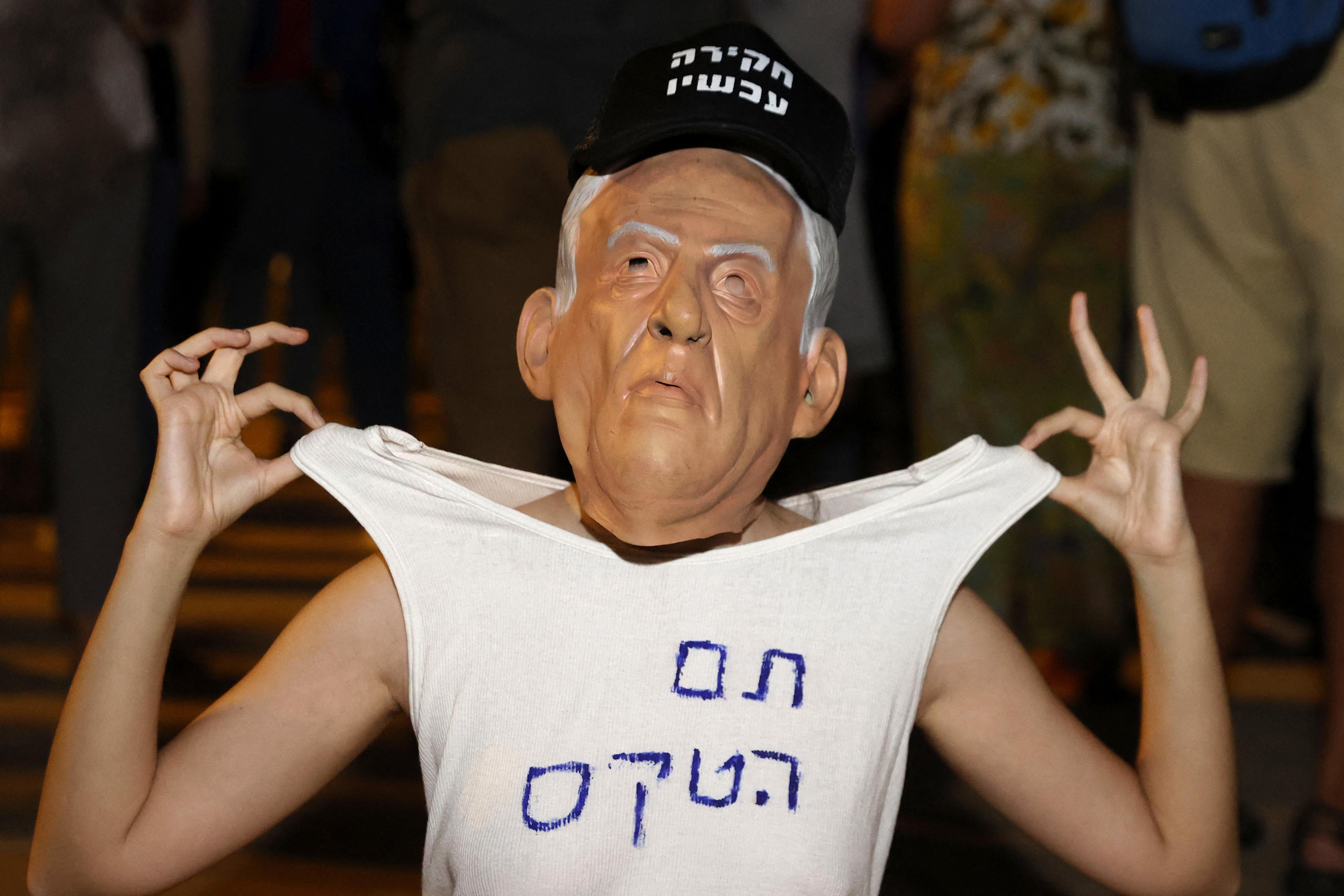 A young woman weras a mask of Benjamin Netanyahu's face, a hat, and a t-shirt with a slogan in Hebrew. She holds the t-shirt up by the shoulders with both hands.
