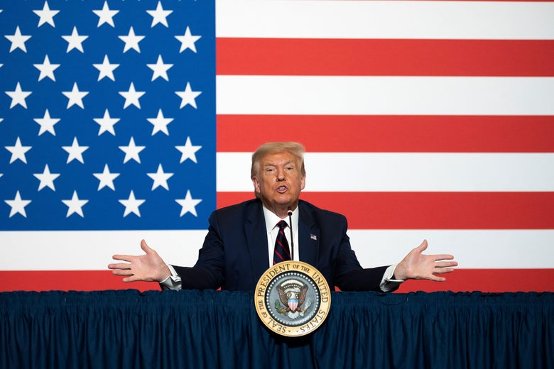 Donald Trump sits in front of an American flag backdrop, raising his hands.