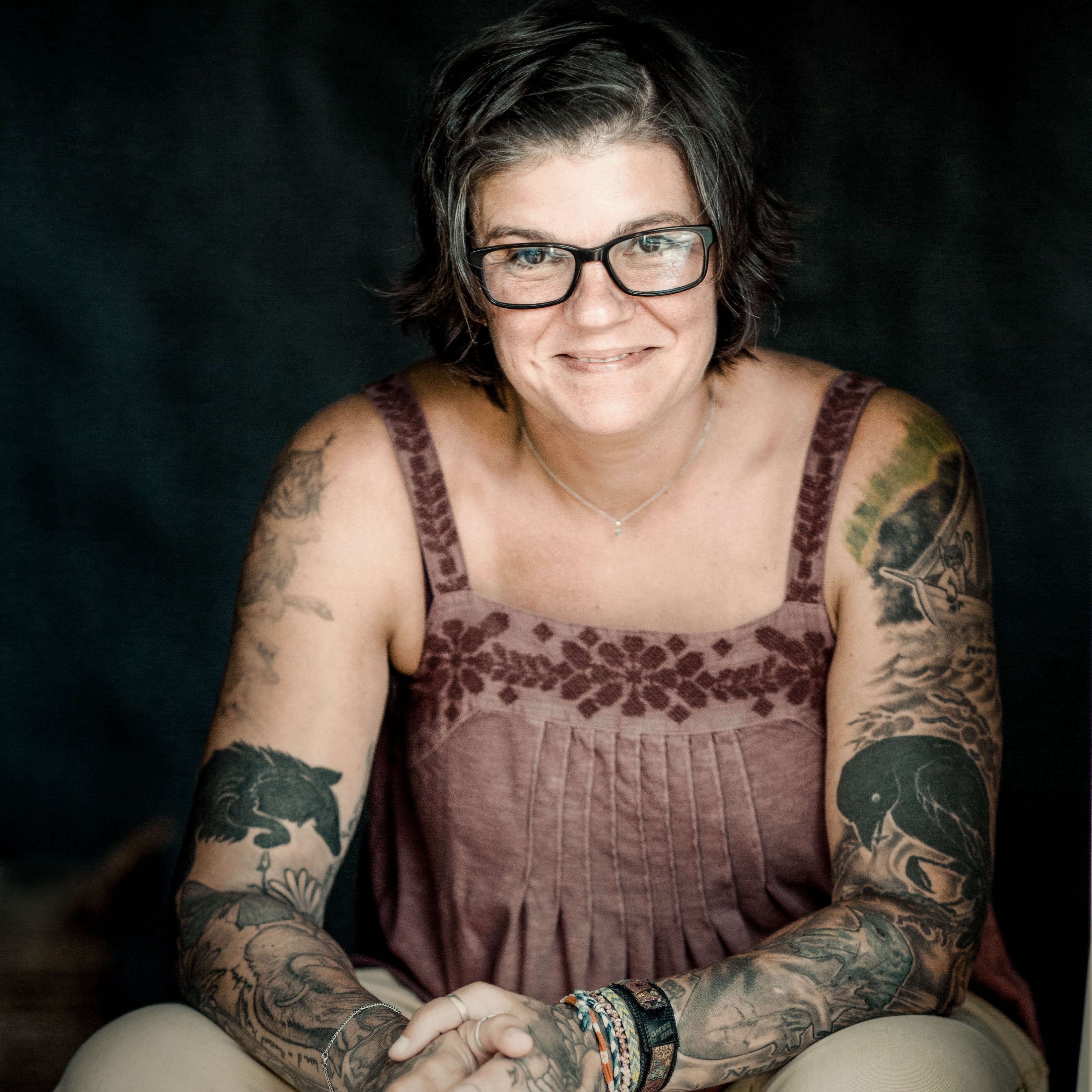 A white woman with short hair and full sleeves of tattoos smiles at the camera.
