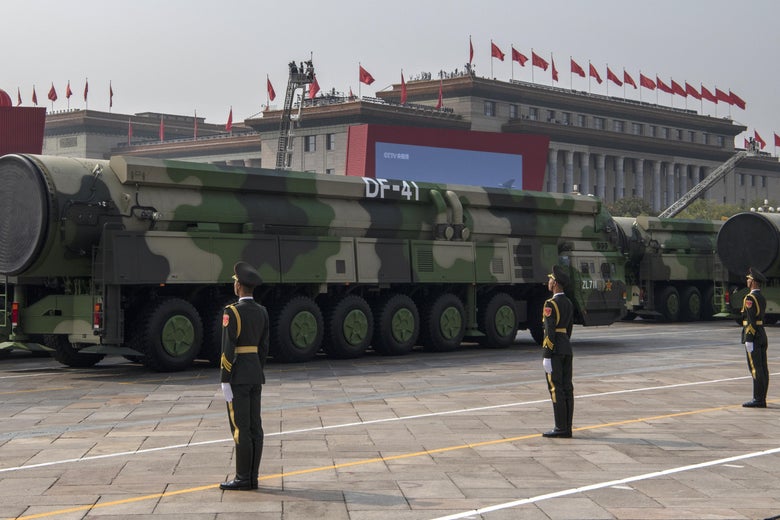 Military vehicles carrying large missiles, with soldiers standing at attention nearby
