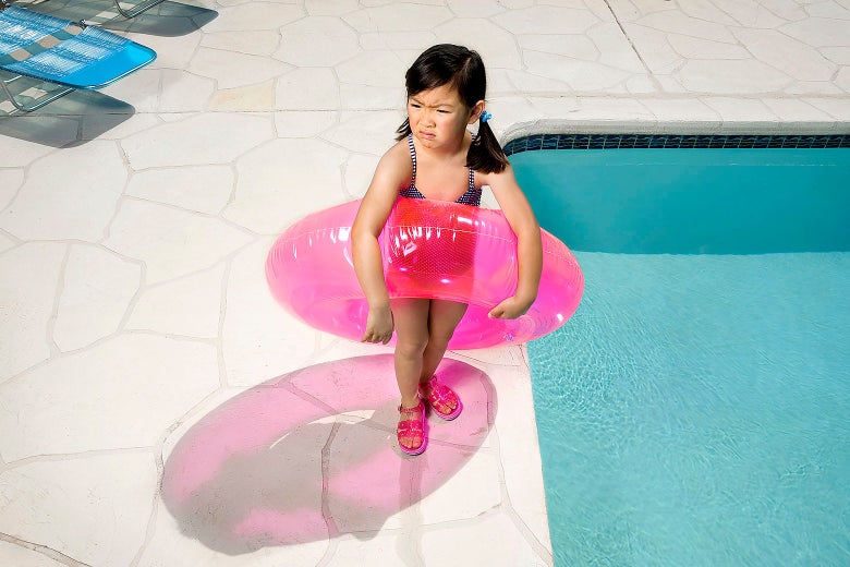A girl in a swimsuit holding a pink inner tube around her waist pouts as she stands next to a pool
