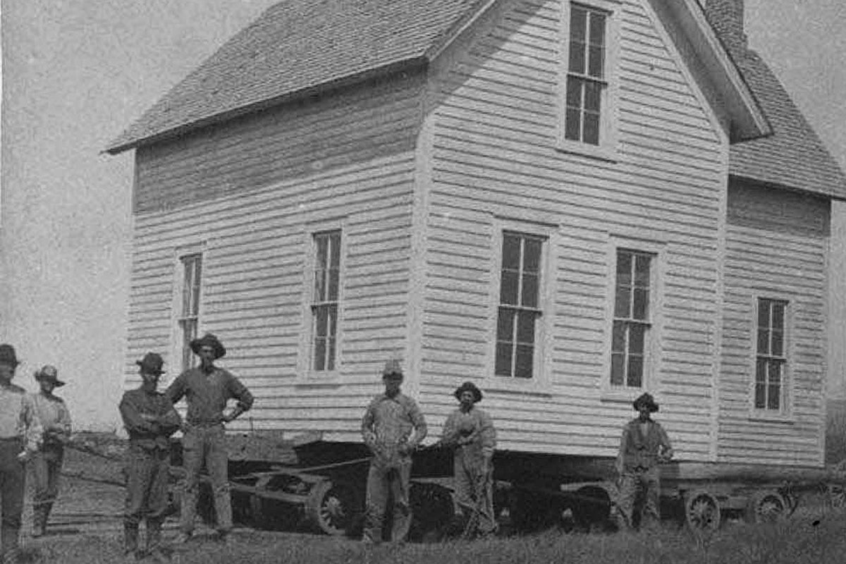 Several man stand beside a two-story house on a large cart.