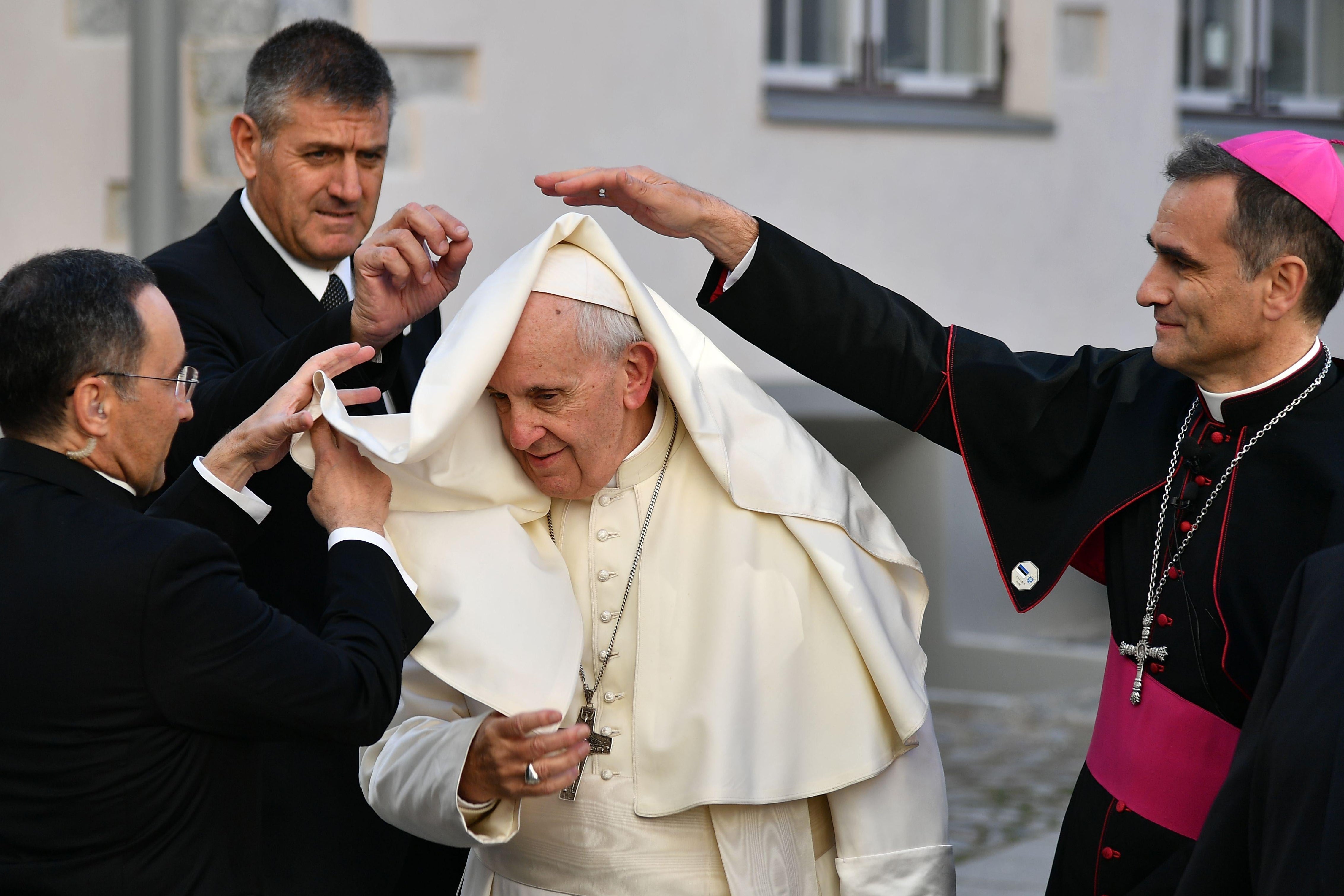 Three people stand around Pope Francis and reach out to pull down his cape, which has blown onto his head.