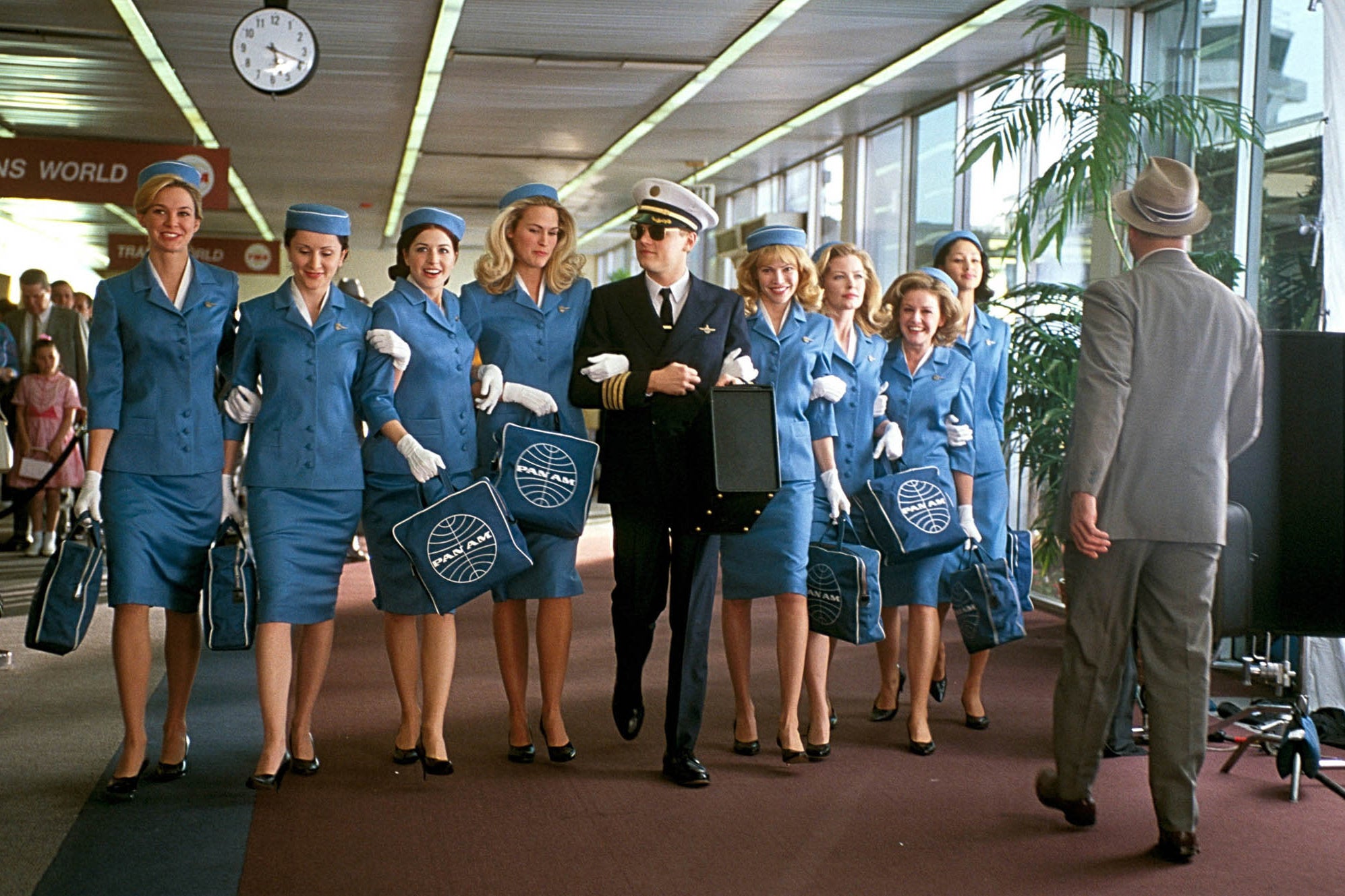 Leonardo DiCaprio dressed as a commercial pilot with sunglasses and a briefcase walks through an airport arm in arm with a line of eight Pan Am flight attendants dressed in cerulean uniforms. Smiles are on everyone's faces. 