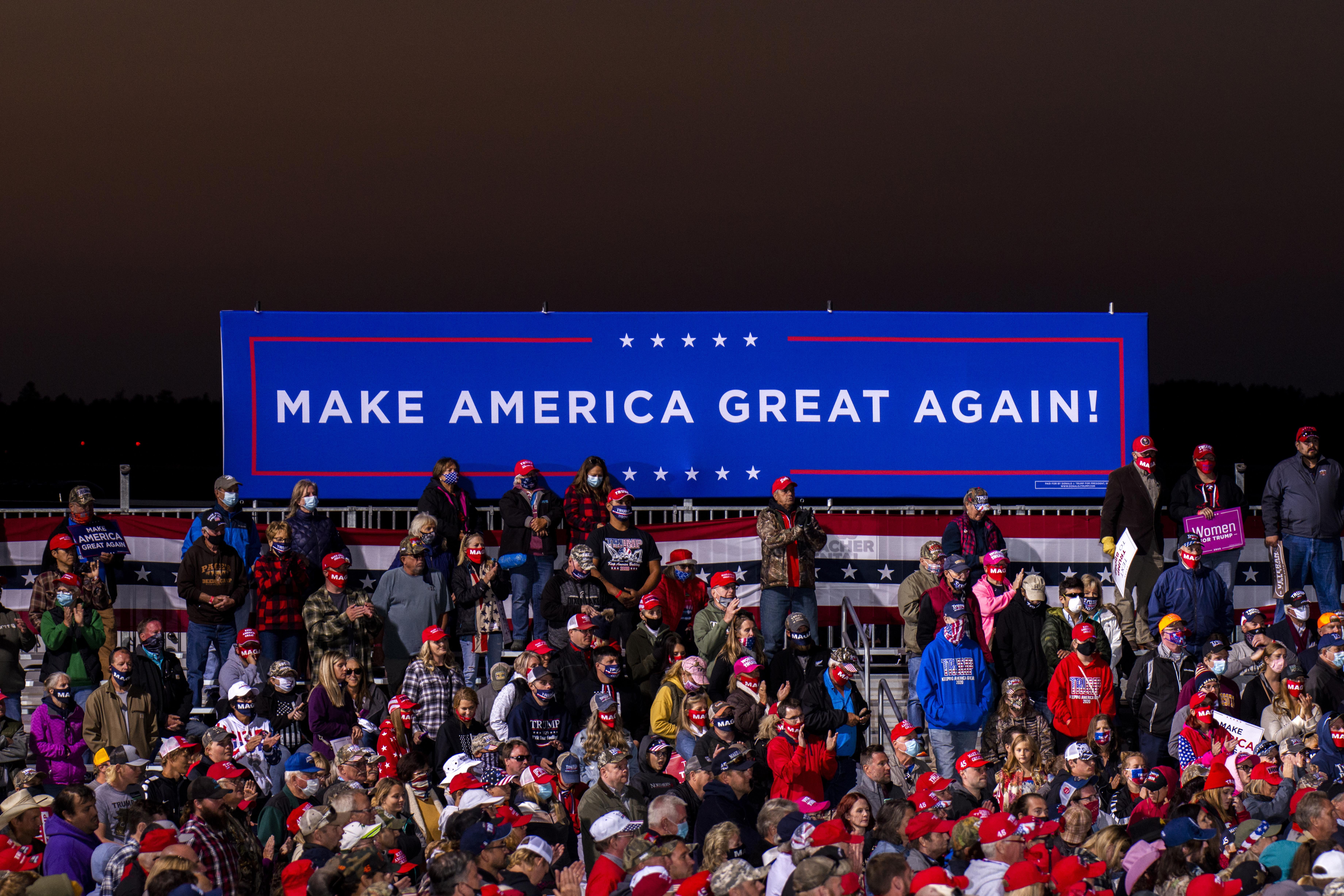 Rows of people, many wearing red caps, sit or stand below a long "Make America Great Again!" sign.