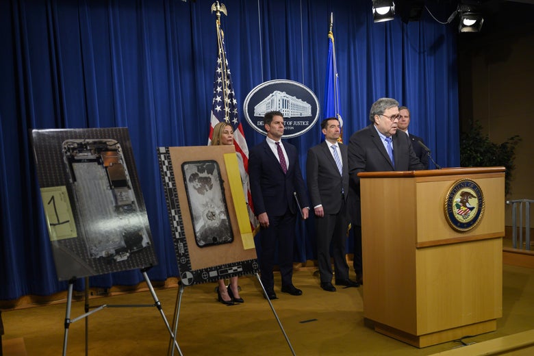 Attorney General William Barr stands at a lectern and speaks, next to pictures of the Pensacola shooter's cellphones.