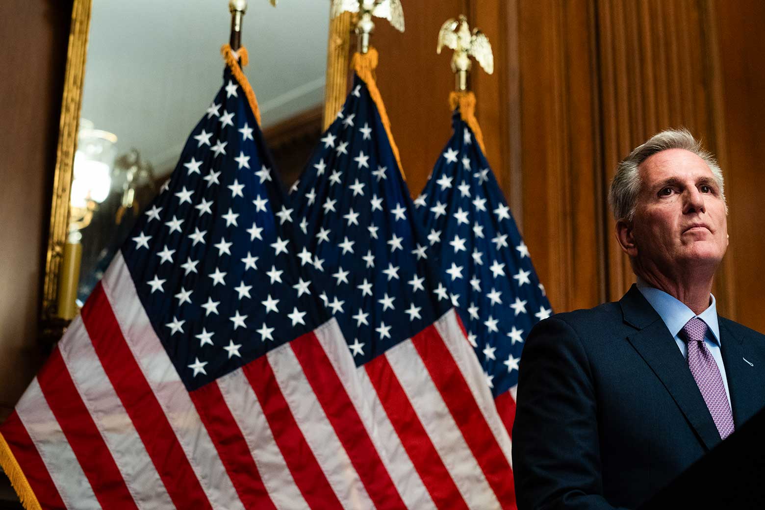 McCarthy looks solemn in front of wood paneling and a series of American flags.