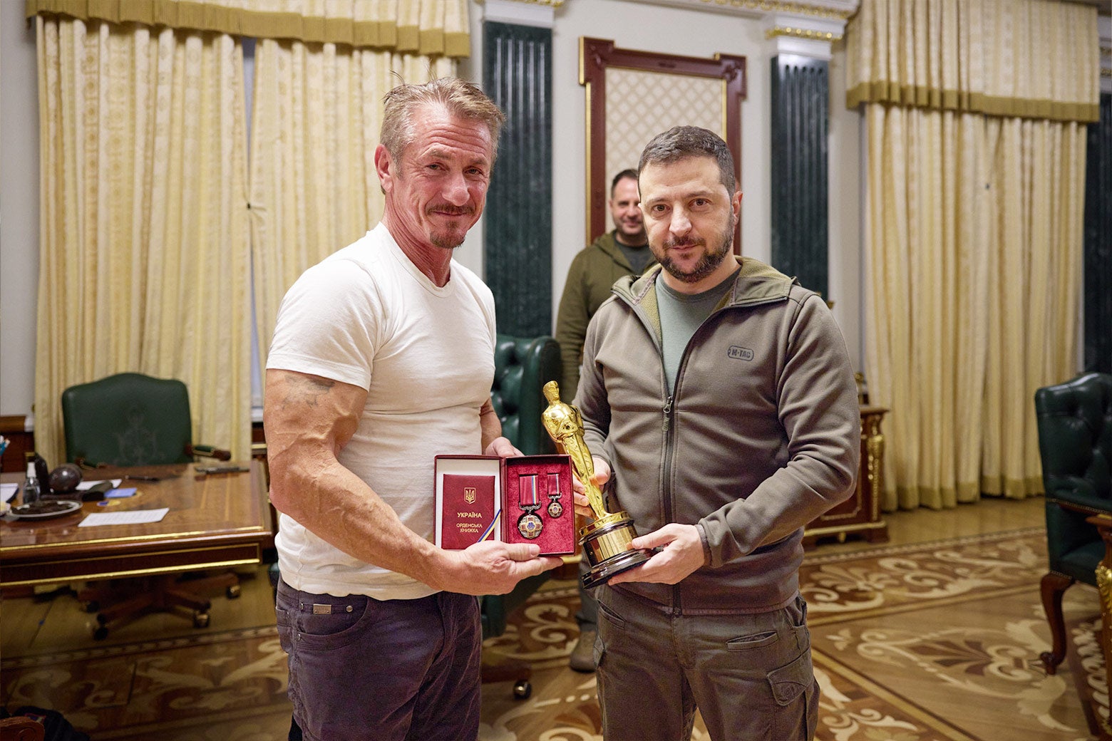 Sean Penn and Volodymyr Zelensky stand next to each other; Zelensky holds Penn's Academy Award and Penn is holding medals. 