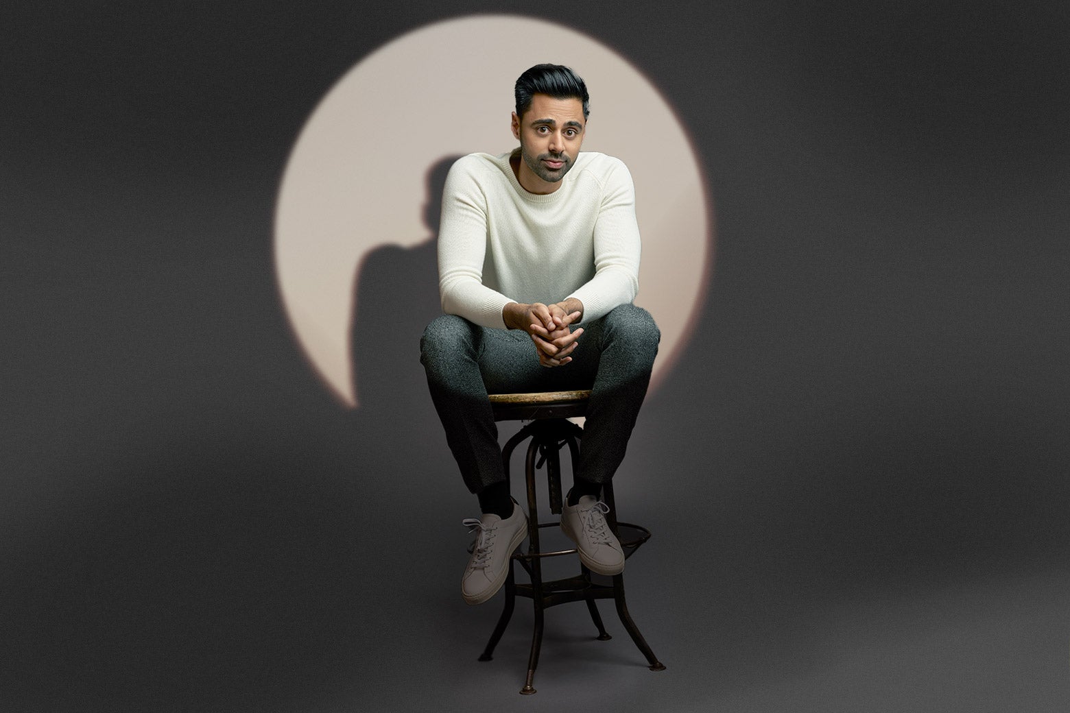 Hasan Minhaj sitting on a stool, with a spotlight on him.