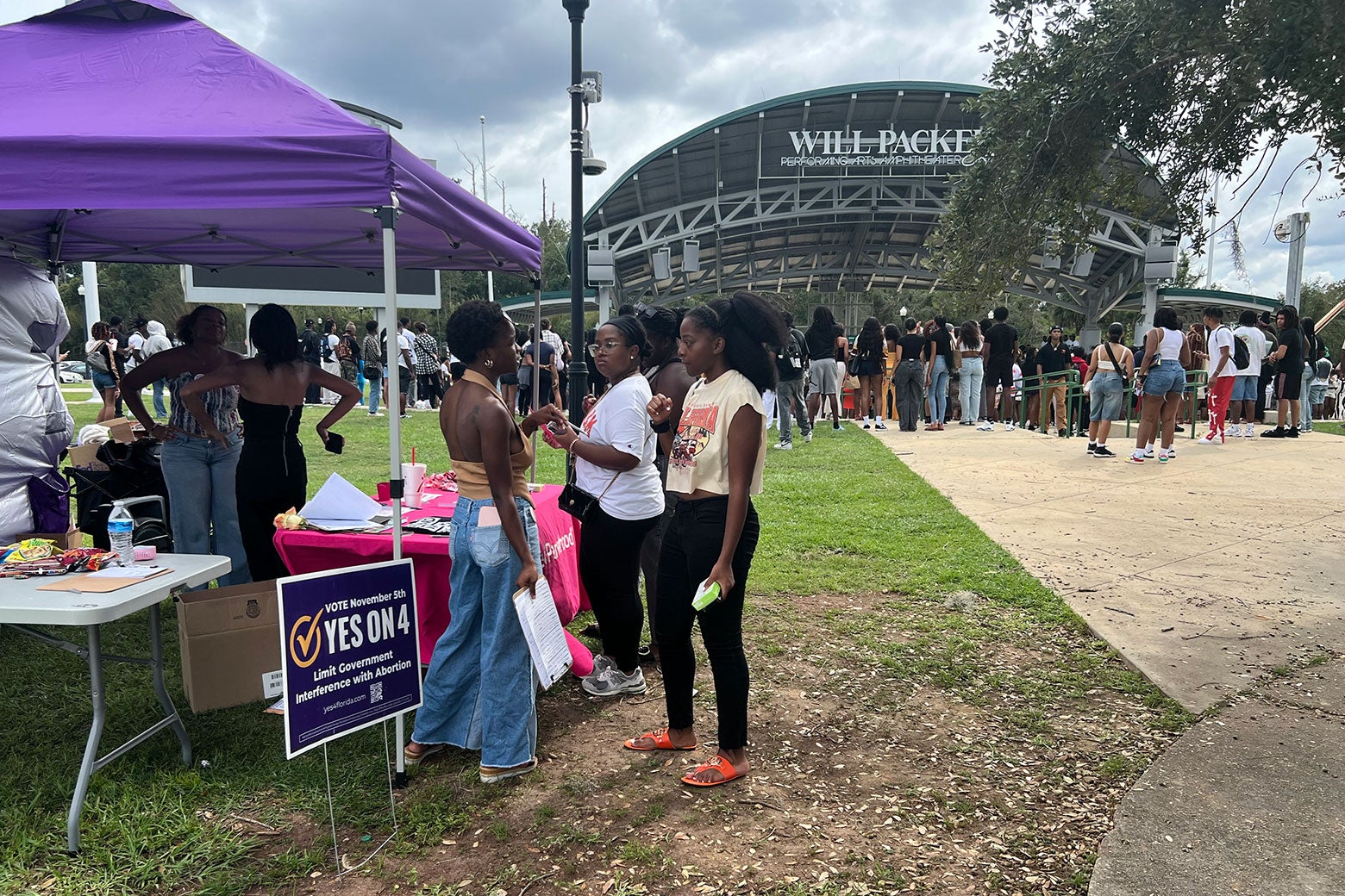 A woman in jeans and a halter top speaks to two other women in front of a purple tent near a sign that says, "Yes on 4."