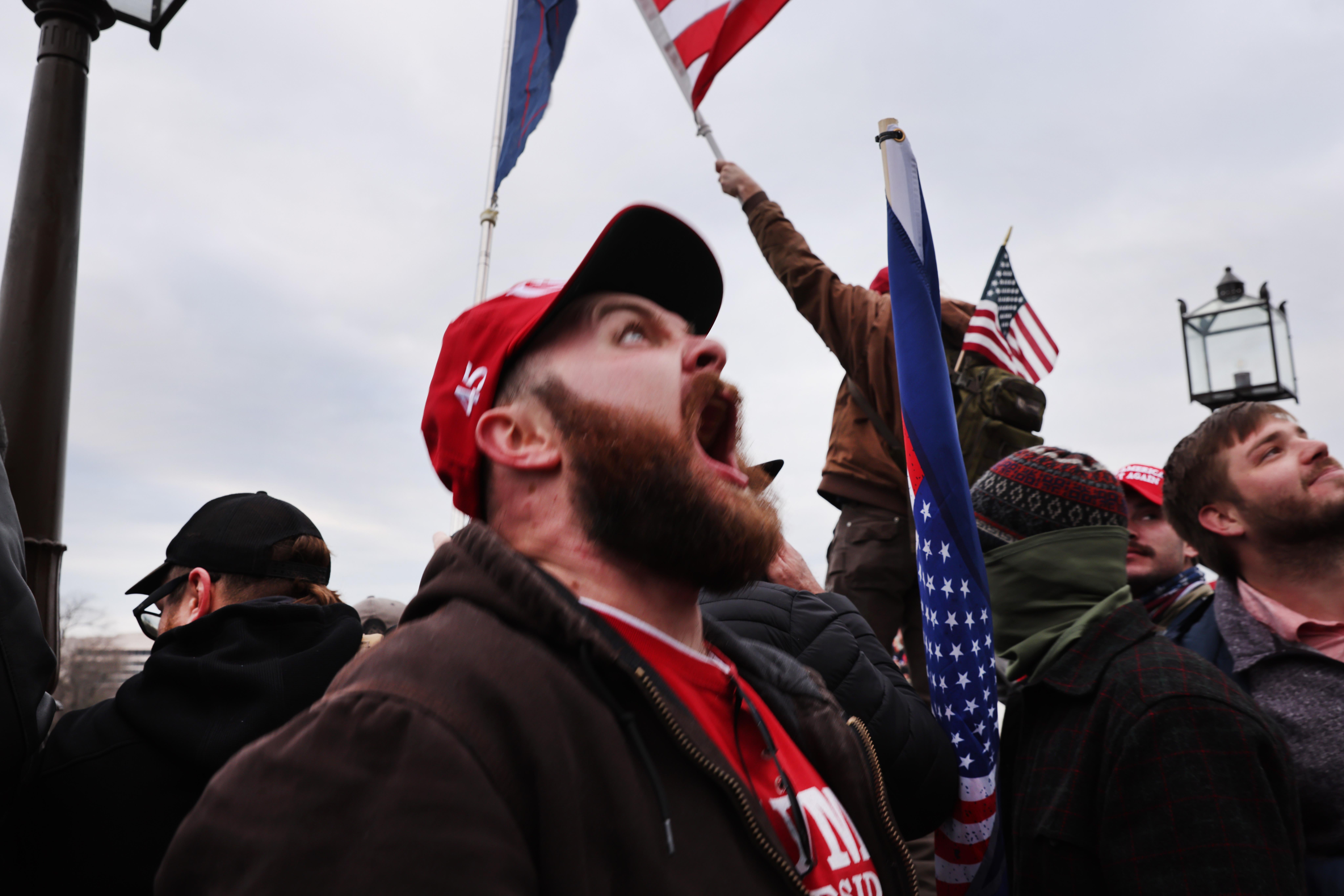 A white man wearing a red 45 hat screams skyward, standing in a crowd of other male Trump supporters, some waving American flags.