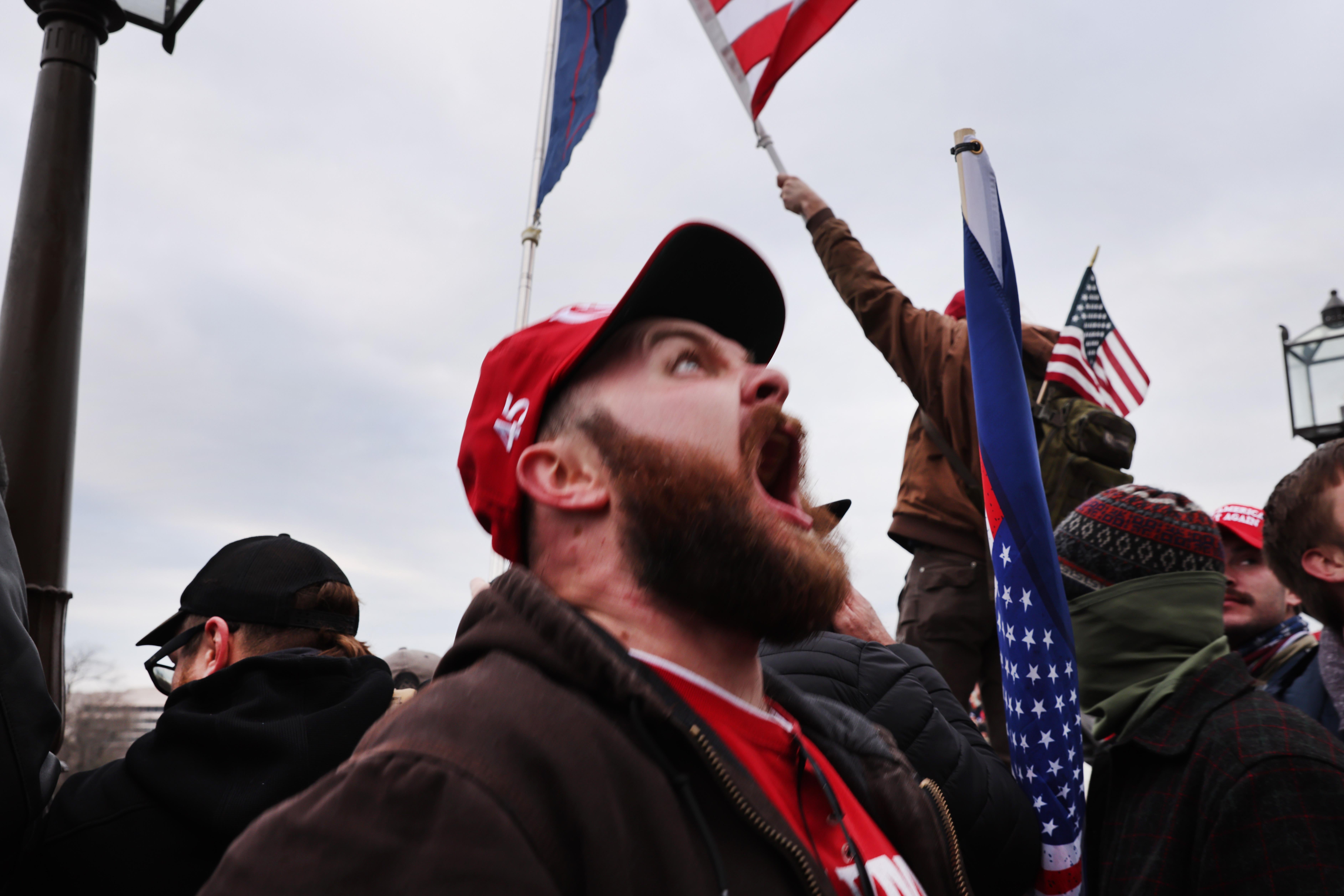 A white man wearing a red 45 hat screams skyward, standing in a crowd of other male Trump supporters, some waving American flags.