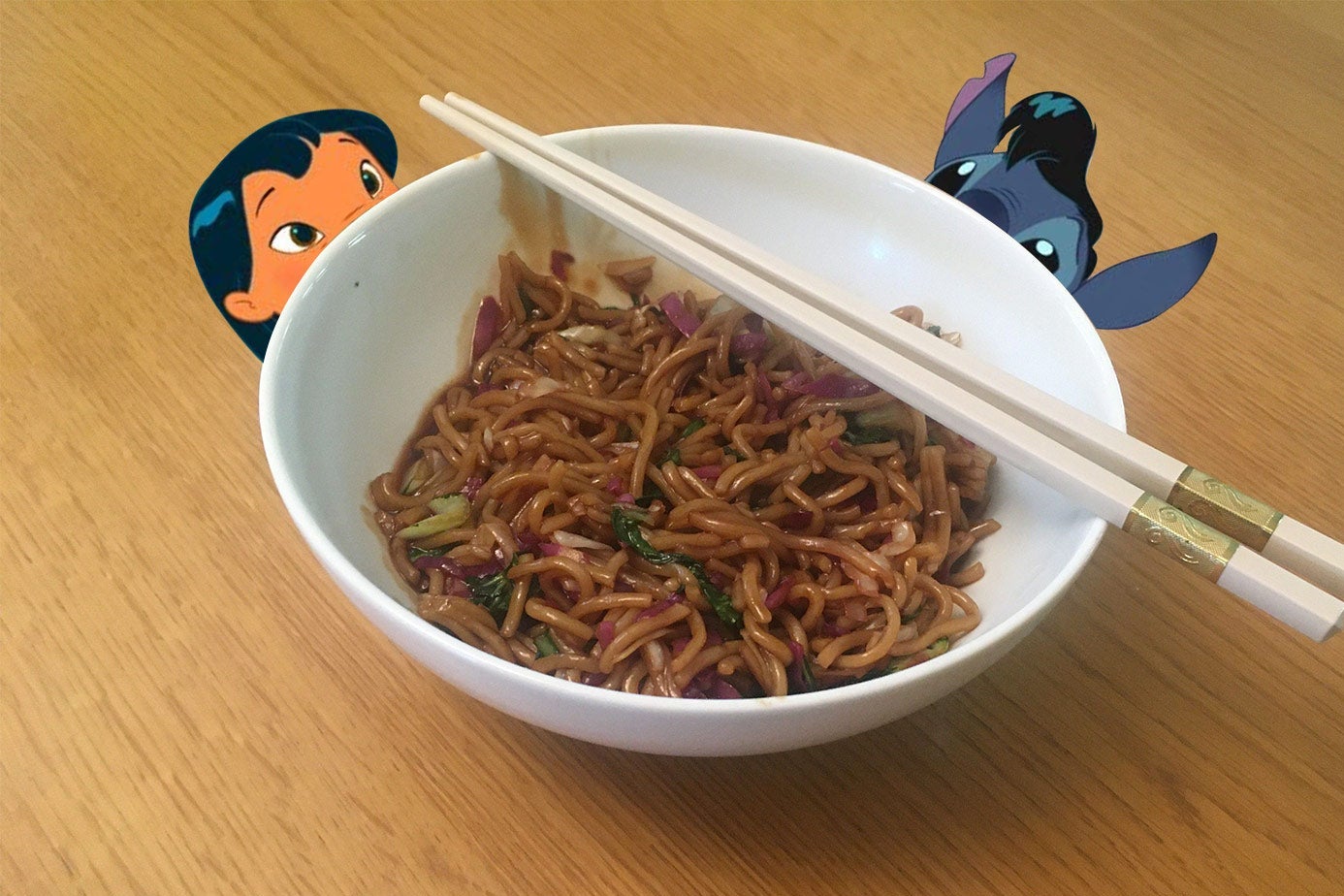 A photo of a bowl of heavily sauced, stir-fried noodles. Peeking over the edge of the bowl are Lilo and Stitch.