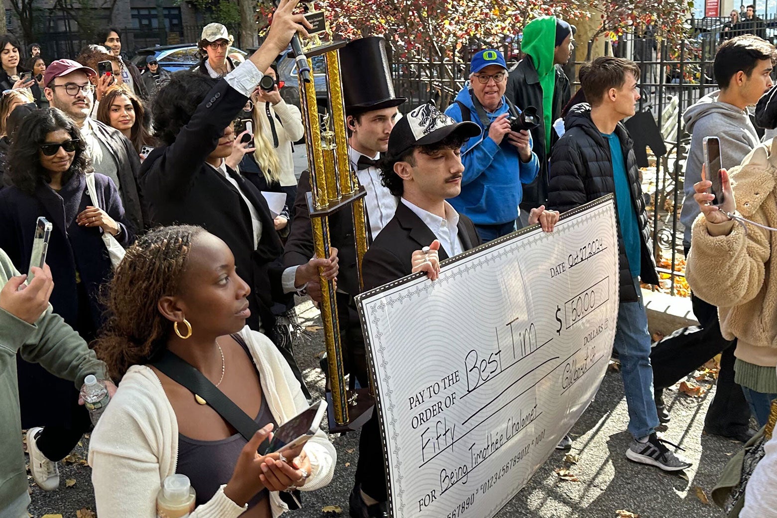 A man in a suit and a black trucker cap holds a huge check for fifty dollars in his hands. "Best Tim." Behind him, event organizer Anthony Poe, wearing a tuxedo and a very tall top hat, holds a large trophy. Two men carry prizes through a crowd of people surrounding them on a city street. 