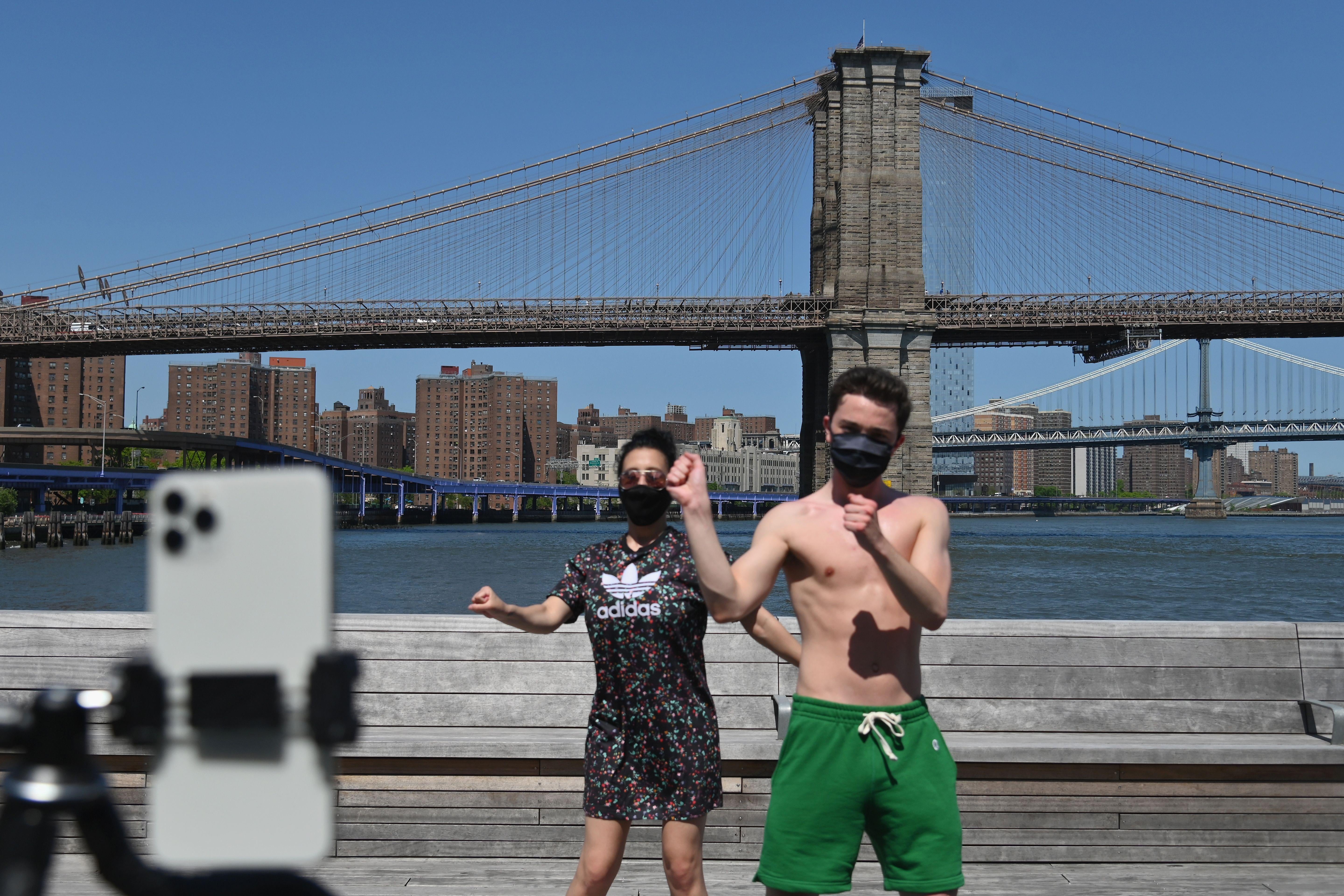 Young man and a woman wearing facemasks dance for a camera phone in front of the Brooklyn Bridge.
