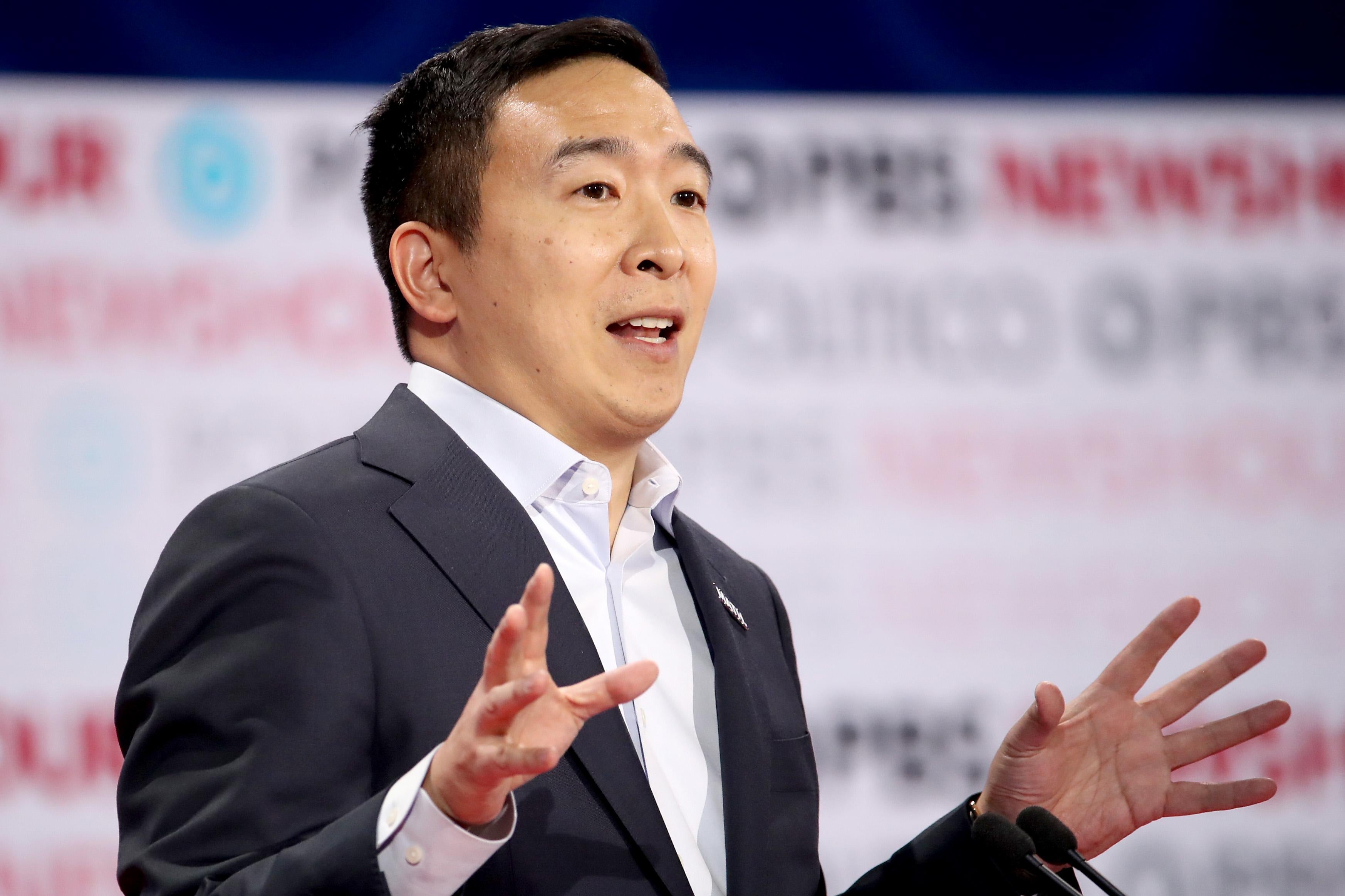 Andrew Yang speaks with raised hands at a podium on the debate stage