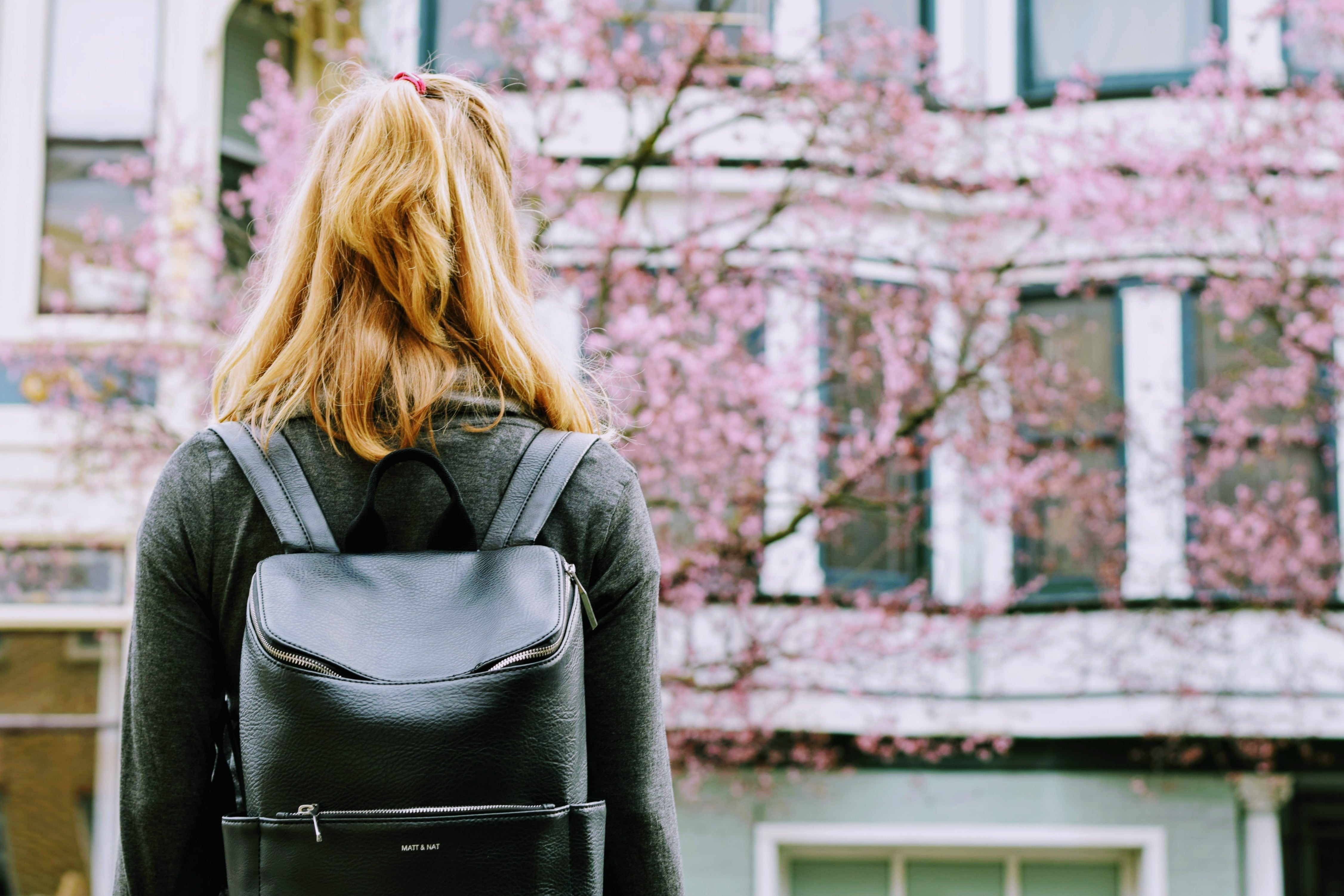The back of a blonde woman with a half pony tail, a long-sleeve shirt, and a black backpack. She looks like she's headed to school. 