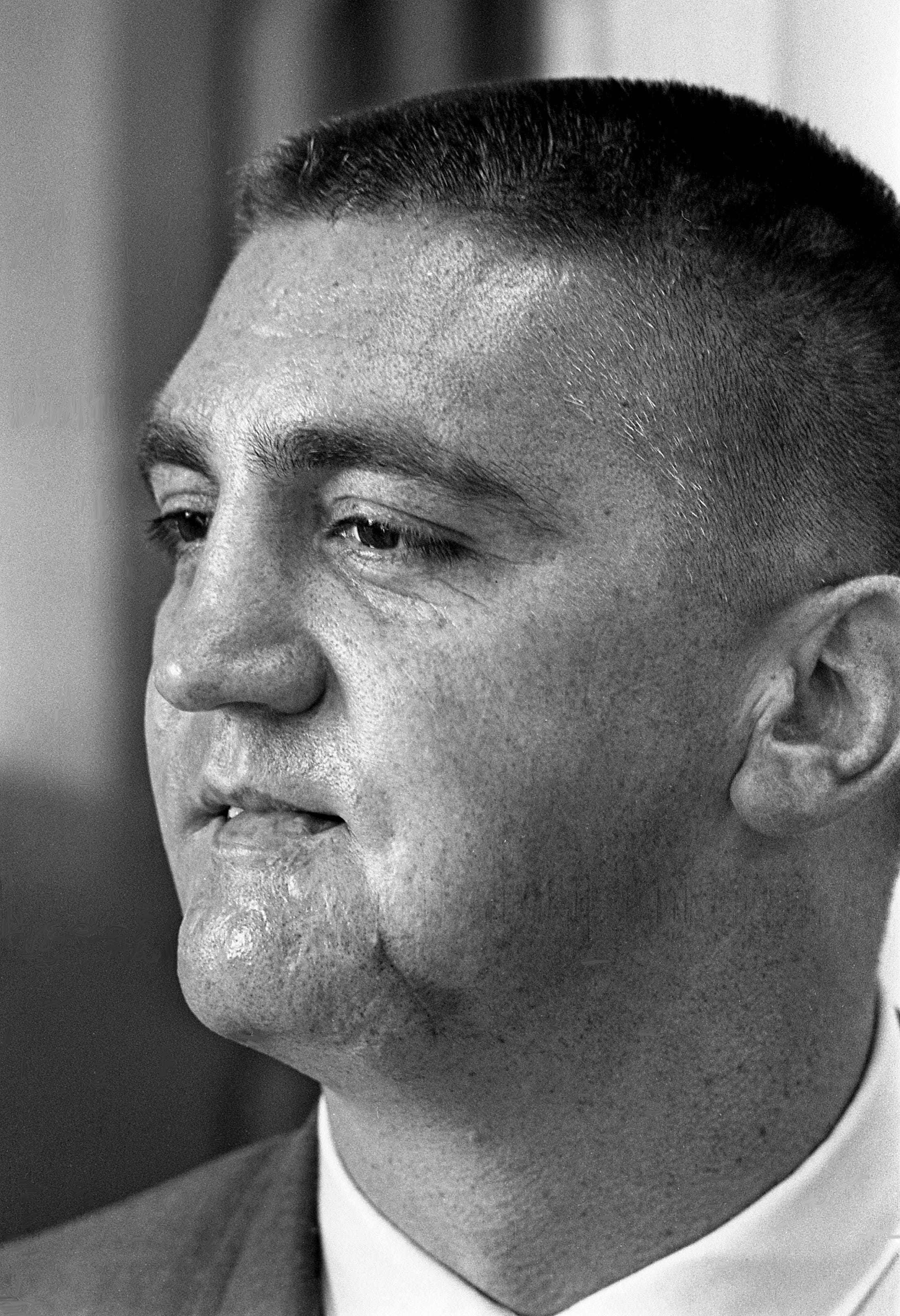 Black-and-white image of a middle-aged man with a buzzcut and a heavily scarred face.