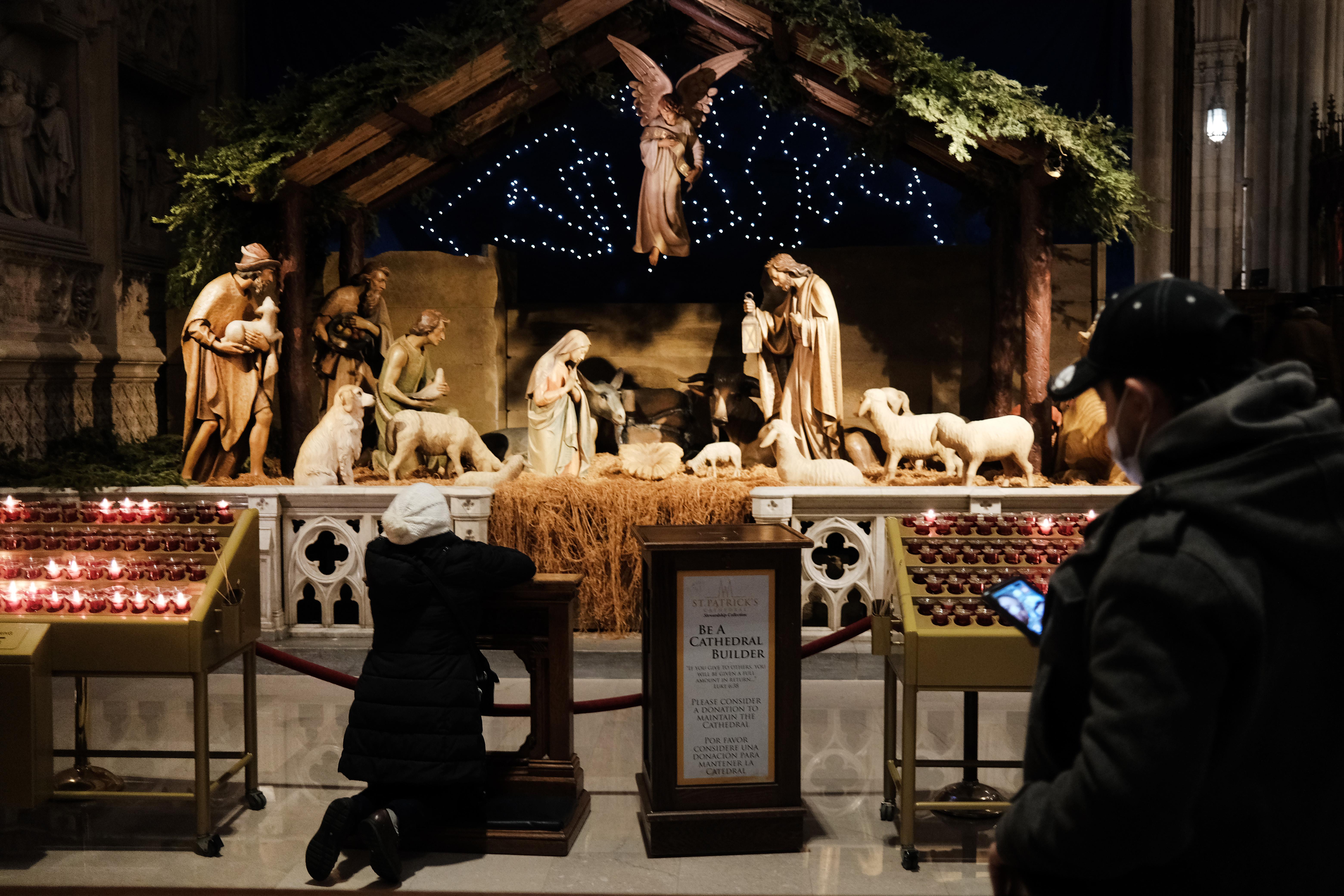 A person kneels in front of a Nativity scene at St. Patrick's Cathedral in New York. Behind her a person wearing a mask stands looking at their phone.