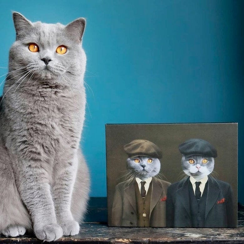 Gray cat sitting next to a small portrait of two gray cats styled as characters from Peaky Blinders wearing tweed suits and newsboy caps
