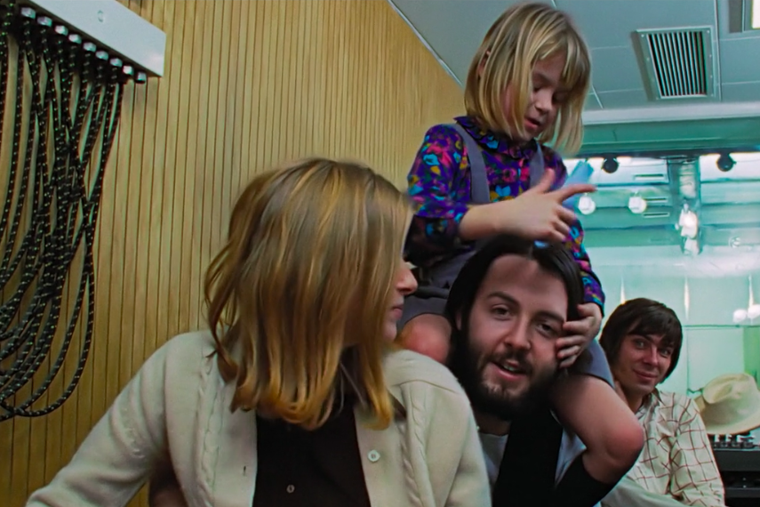 A woman with shoulder-length brown hair has her head turned to the side beside Paul McCartney who is smiling with a little girl sitting on his shoulders