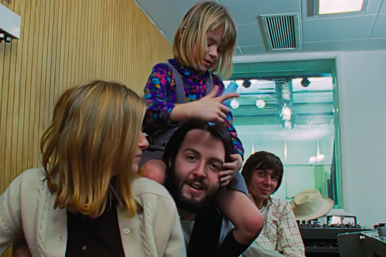 A woman with shoulder-length brown hair has her head turned to the side beside Paul McCartney who is smiling with a little girl sitting on his shoulders