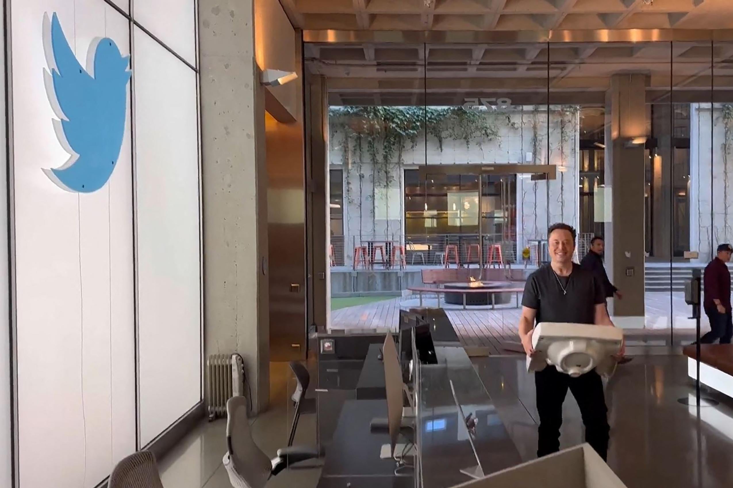 Elon Musk carrying a sink in an office where the Twitter logo is displayed on a wall