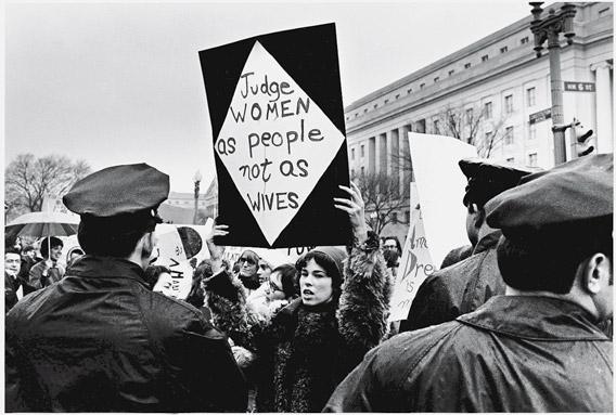 A young American woman holds up a sign as she protests for women's rights in front of the Federal Trade Commission headquarters while policemen look on during Richard Nixon's inauguration weekend, Washington, DC, January 18-21, 1969. Her sign reads 'Judge women as people not as wives.