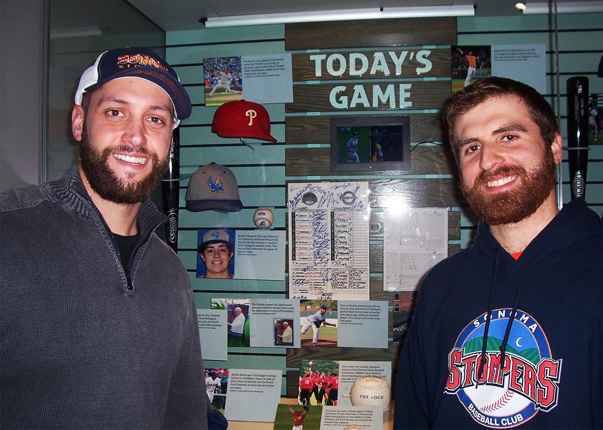 Sean Conroy and Isaac Wenrich pose in Cooperstown in front of a Hall of Fame display case containing the scorecard from the Stompers' Pride Night game. 