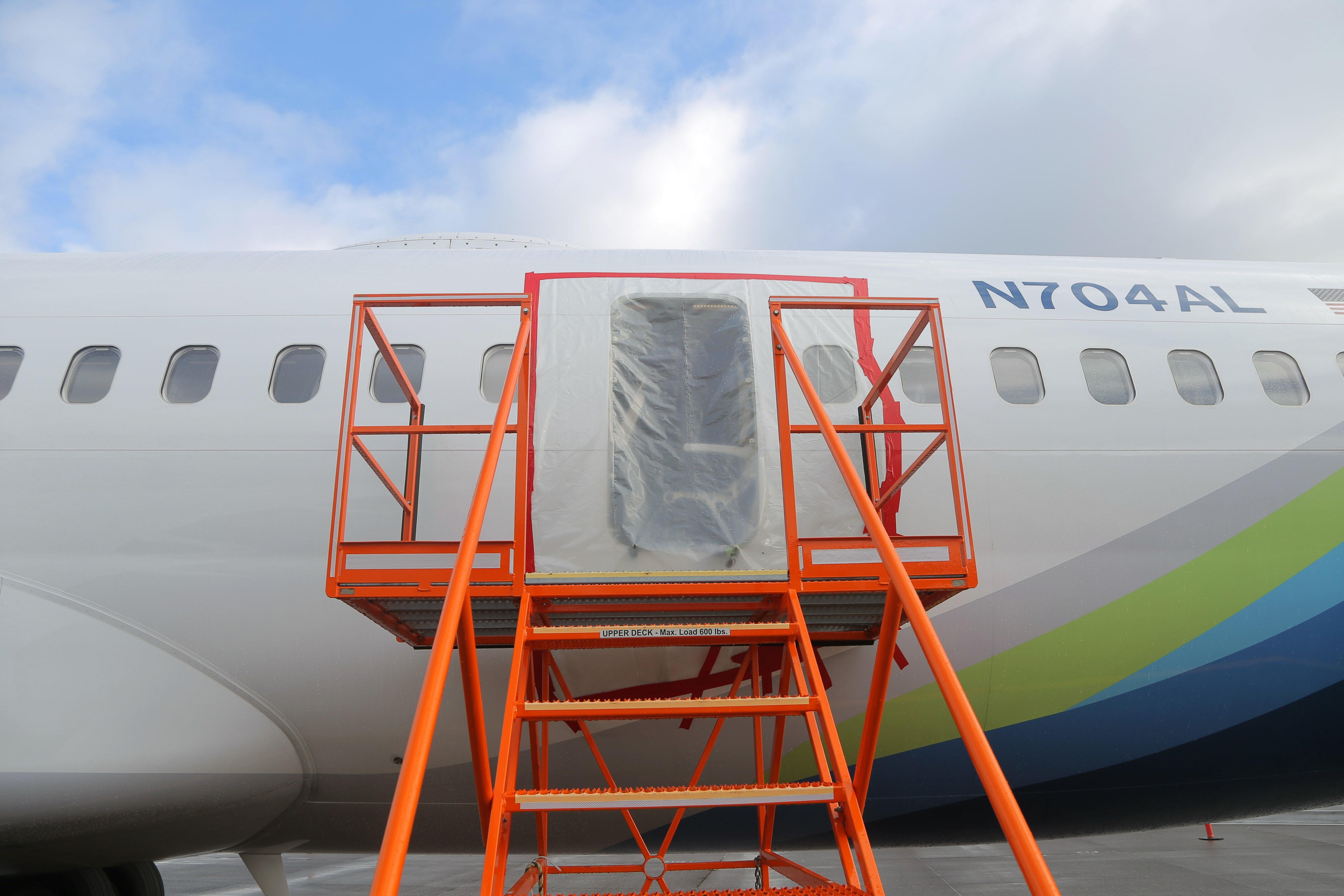 The side of a Boeing 737-9 Max jet from the Alaska Airlines fleet. The space where the door plug should be is covered with a sheet of plastic, and orange stairs lead to the opening. 