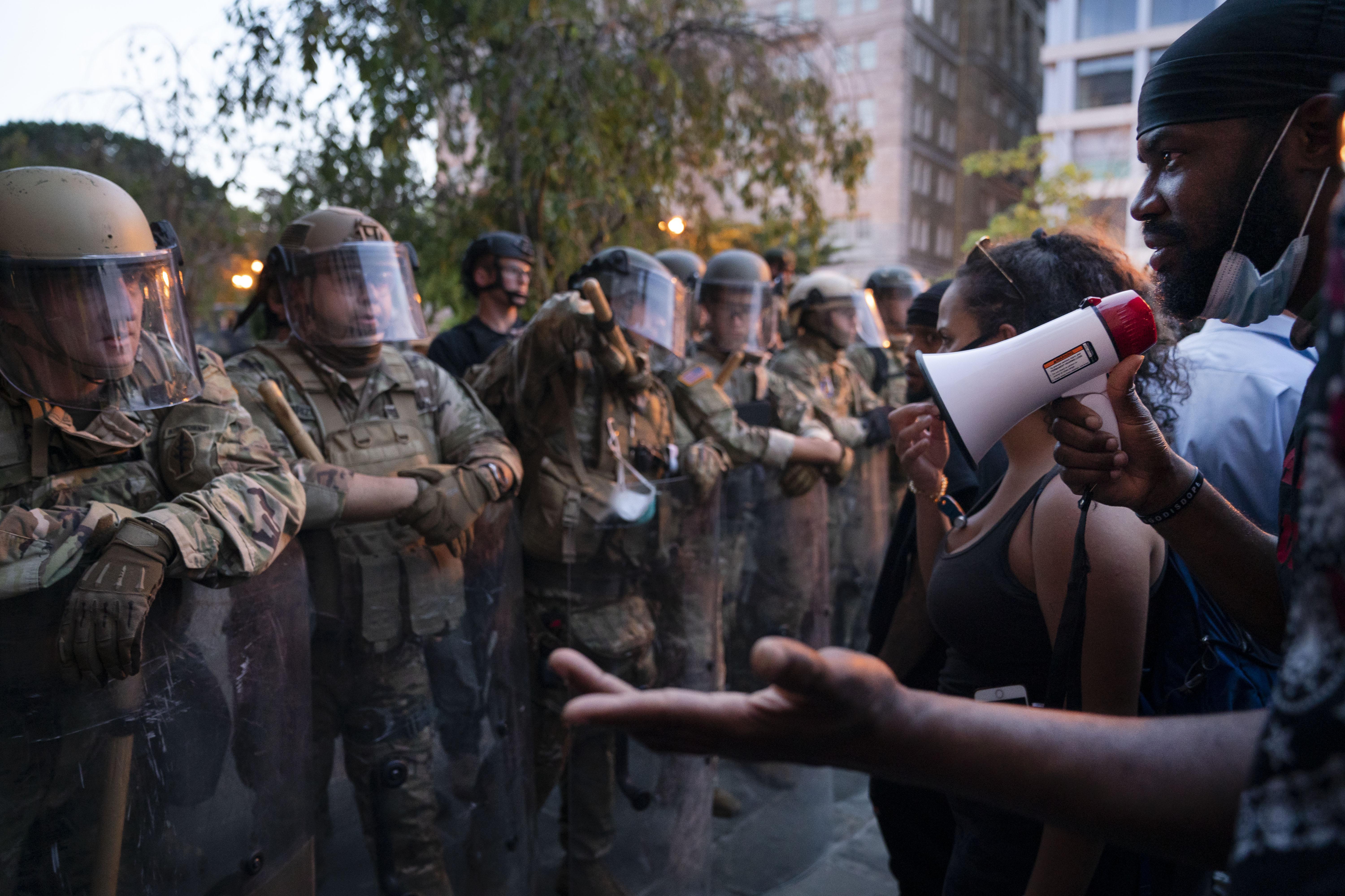 A protester with a bullhorn talks with a line of armed militarized police in camo and riot gear.