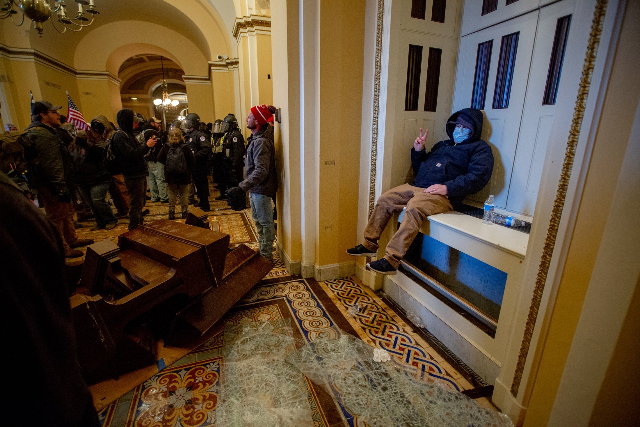 A man sits on a ledge amid a crowd at the Capitol, flashing a peace sign. 