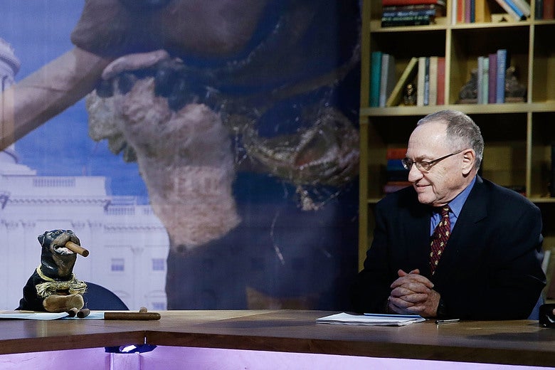 Dershowitz sits at a desk on stage next to a small dog puppet that has a cigar in its mouth.