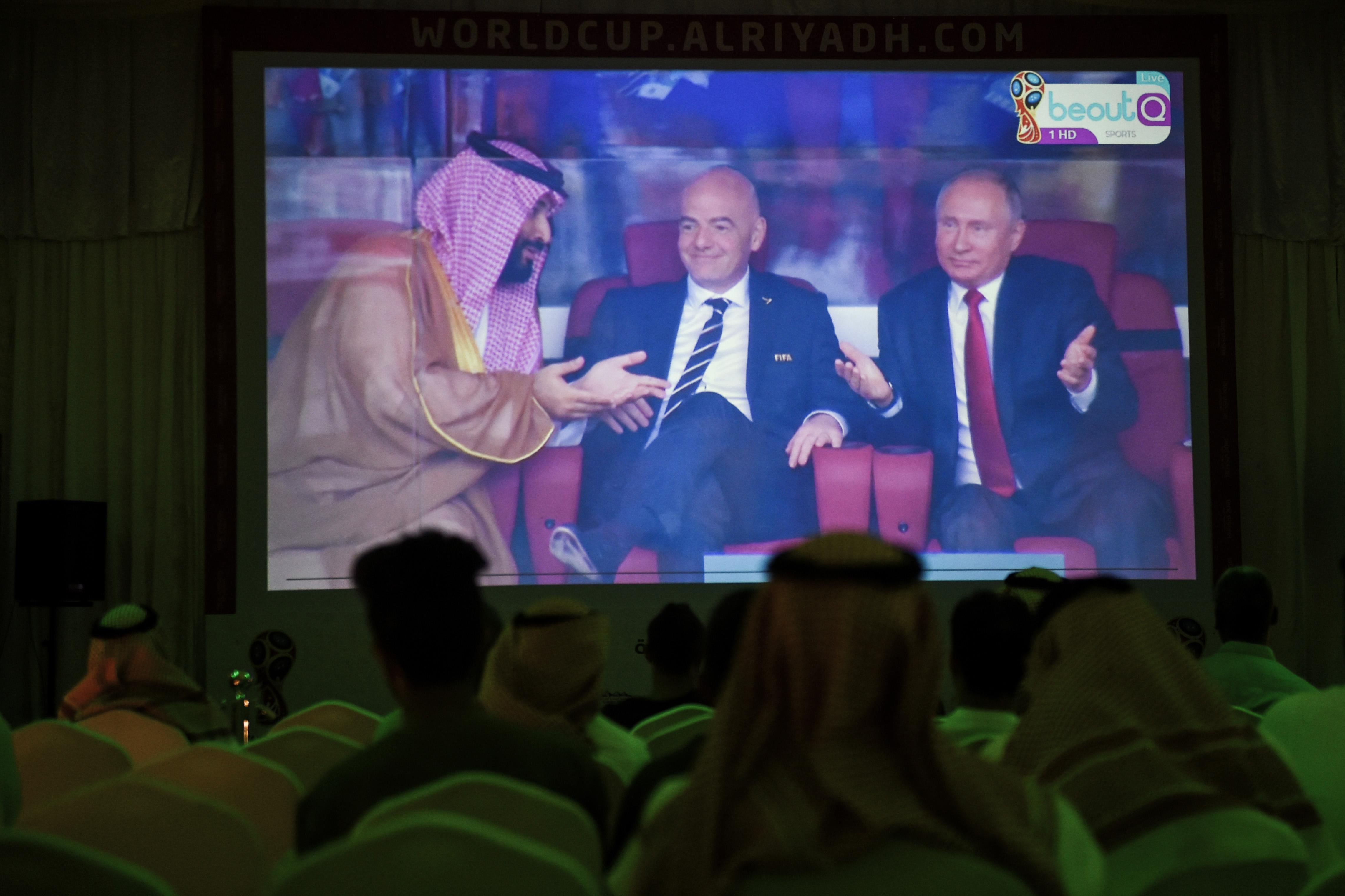 A picture taken on June 14, 2018 in a Saudi football fan tent in the capital Riyadh shows a projector showing the Russia 2018 World Cup Group A football match between Russia and Saudi Arabia, with a clip of Saudi Crown Prince Mohammed bin Salman (L) gesturing as he sits next to FIFA President Gianni Infantino (C) and Russian President Vladimir Putin (R). (Photo by Fayez Nureldine / AFP)        (Photo credit should read FAYEZ NURELDINE/AFP/Getty Images)