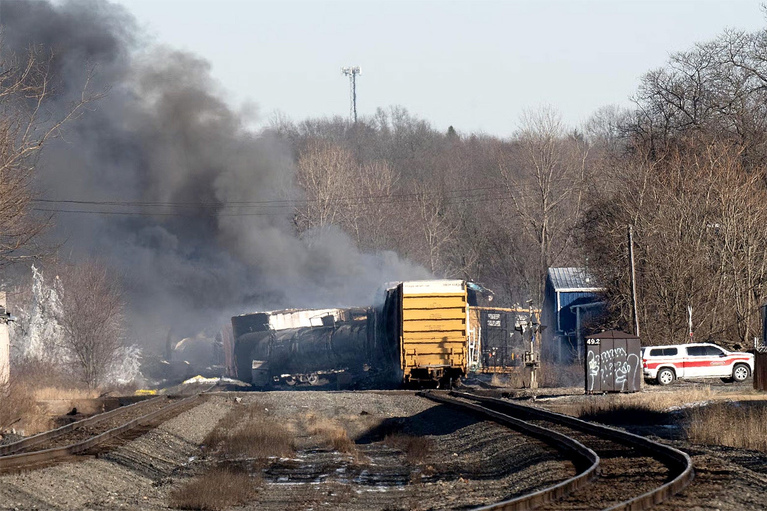 Two train tracks cut through the center of the photo, and black smoke rises from a derailed cargo train, laying on its side across the tracks.