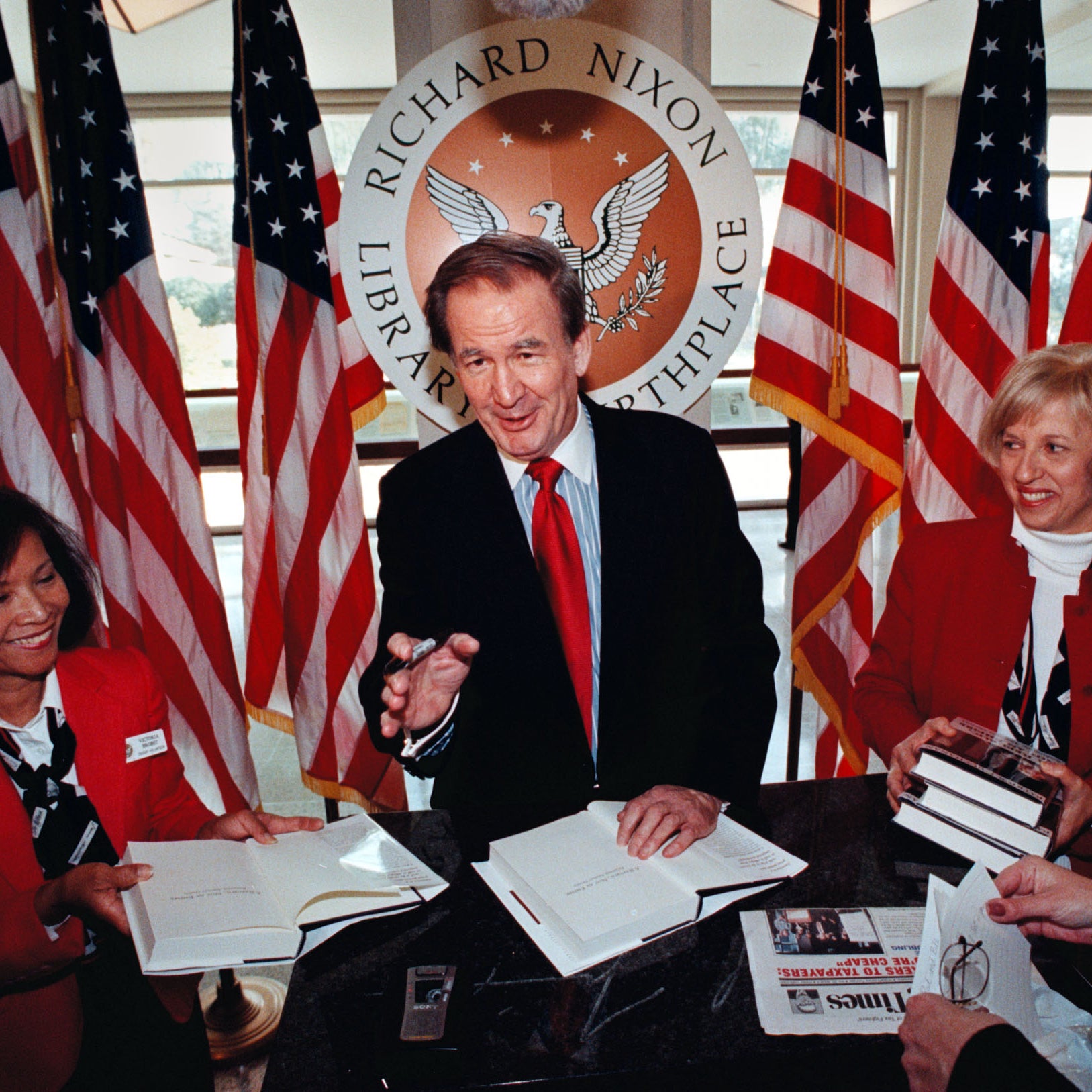 Buchanan speaks while signing books in front of American flags and a Nixon library and birthplace seal, two women in red blazers flanking him, holding books and smiling