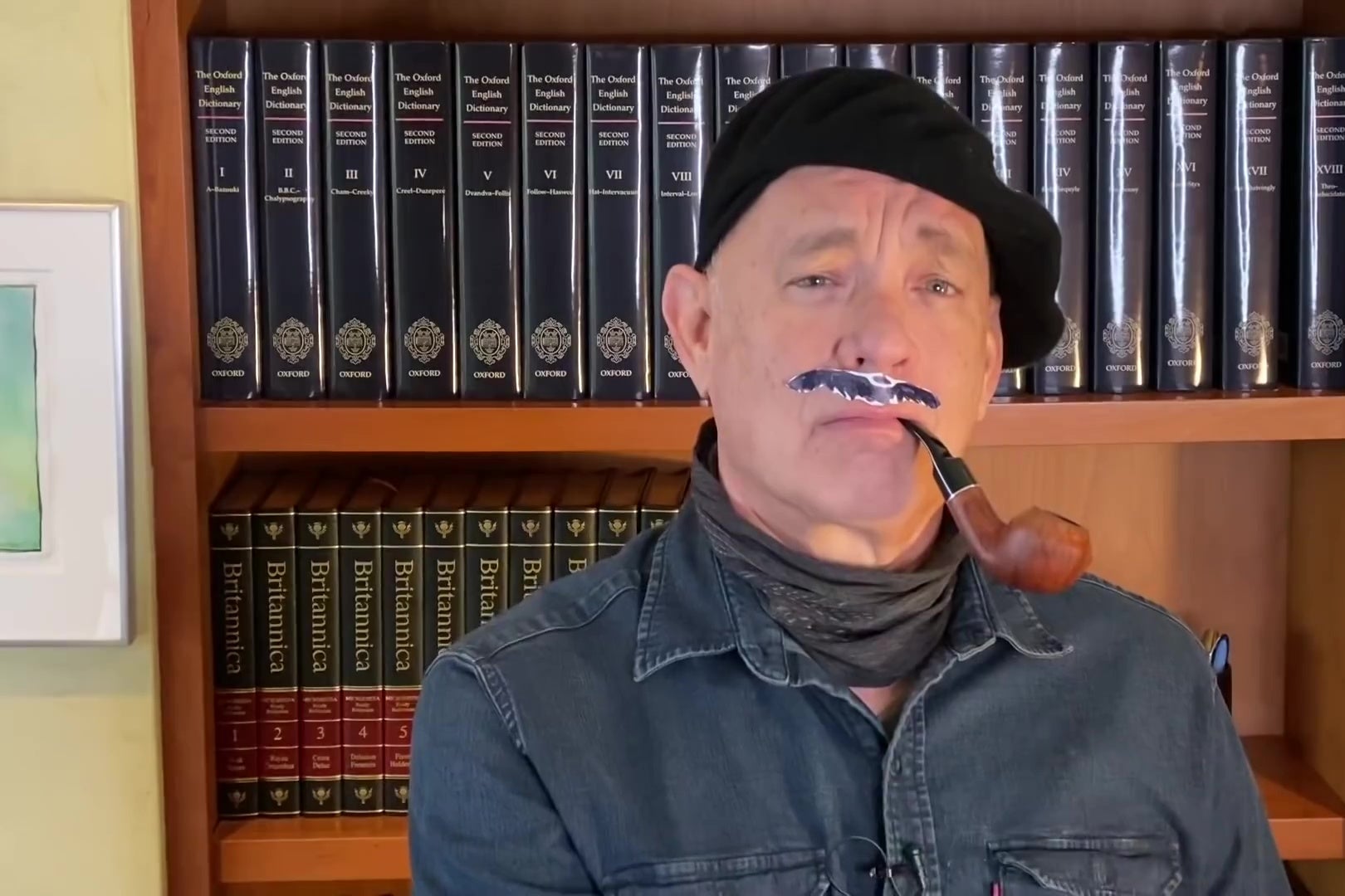 Tom Hanks, wearing a beret, fake mustache, denim shirt, and scarf, smokes a pipe in front of a bookshelf containing the first 18 volumes of the OED and an encyclopedia.