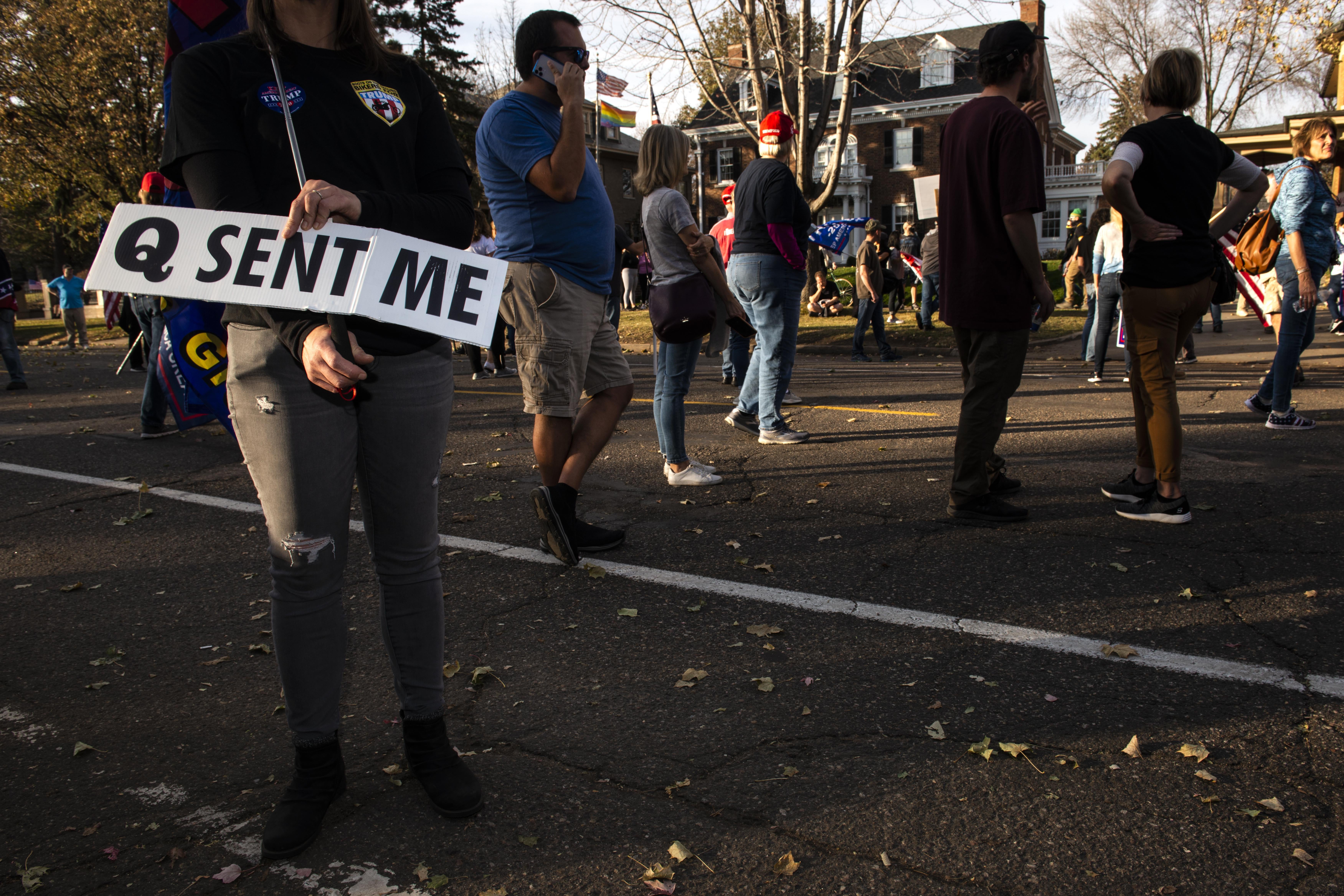 A woman holds a sign reading "Q sent me" on the street at a rally.