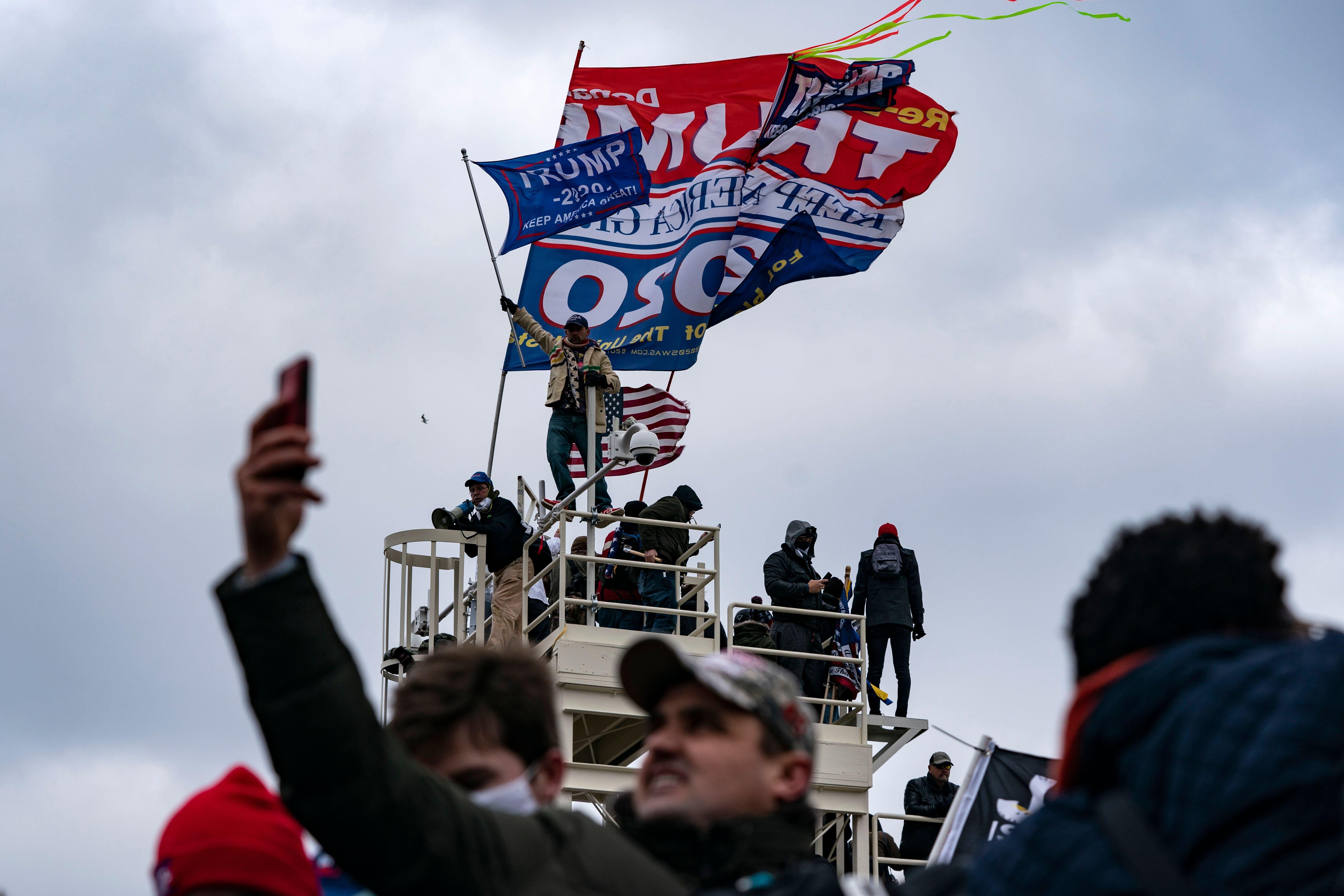 People waving Trump flags mass on top of a platform on a cloudy day. Below, a Trump supporter holds his phone up to take a photo of the chaos.