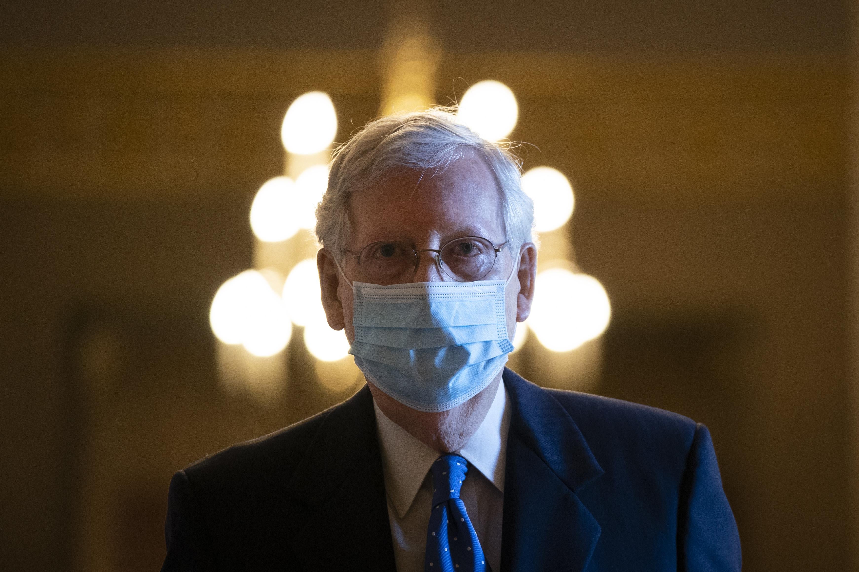 Mitch McConnell walking, wearing a mask, with a chandelier behind his head