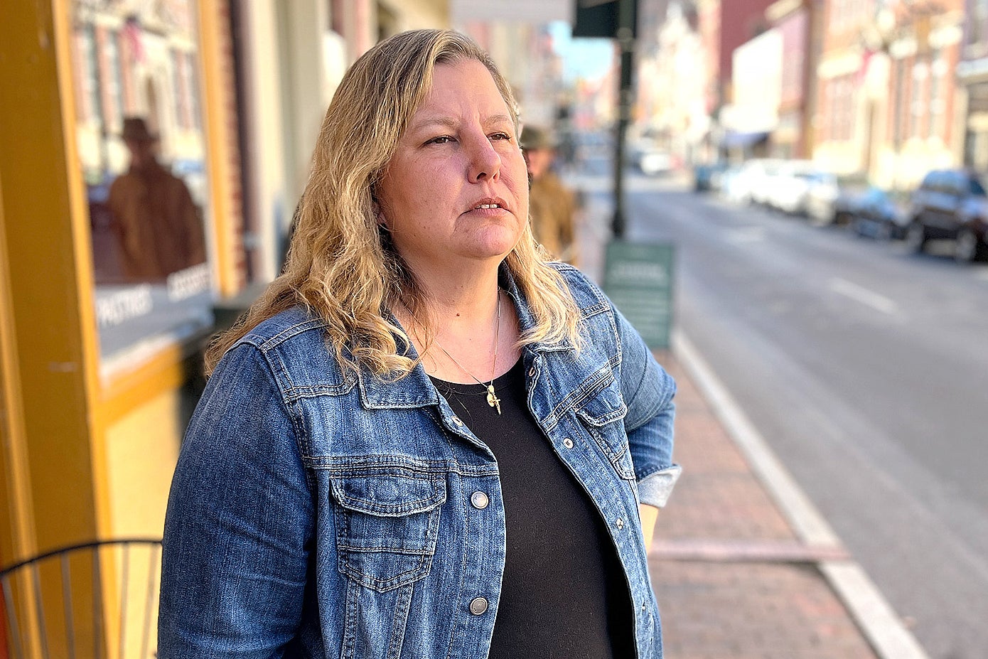 A woman with blond hair, wearing a jean jacket, stands on a sidewalk on a small town downtown street.