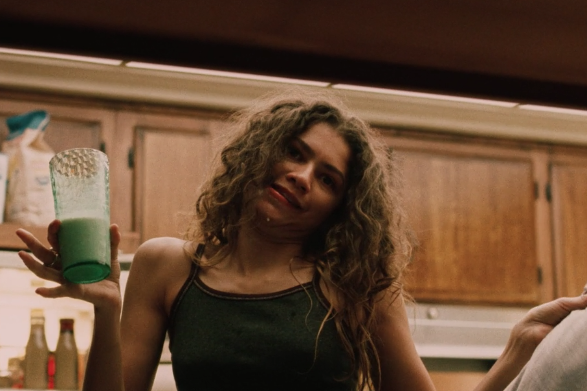 A girl stands in the kitchen holding a pillow and glass of milk.