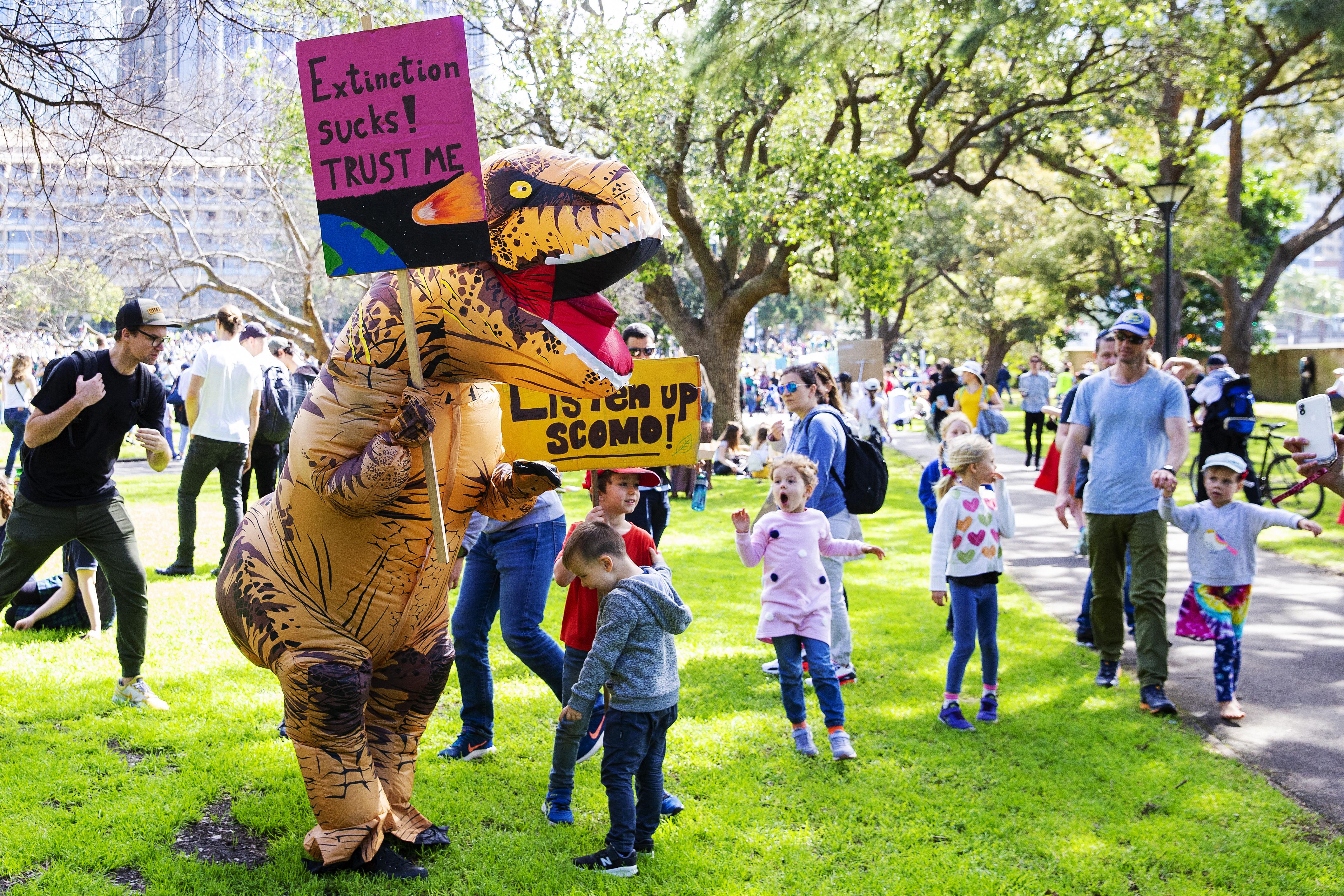 Children react as a man dressed as a dinosaur carries a banner that says "extinciton sucks! trust me"