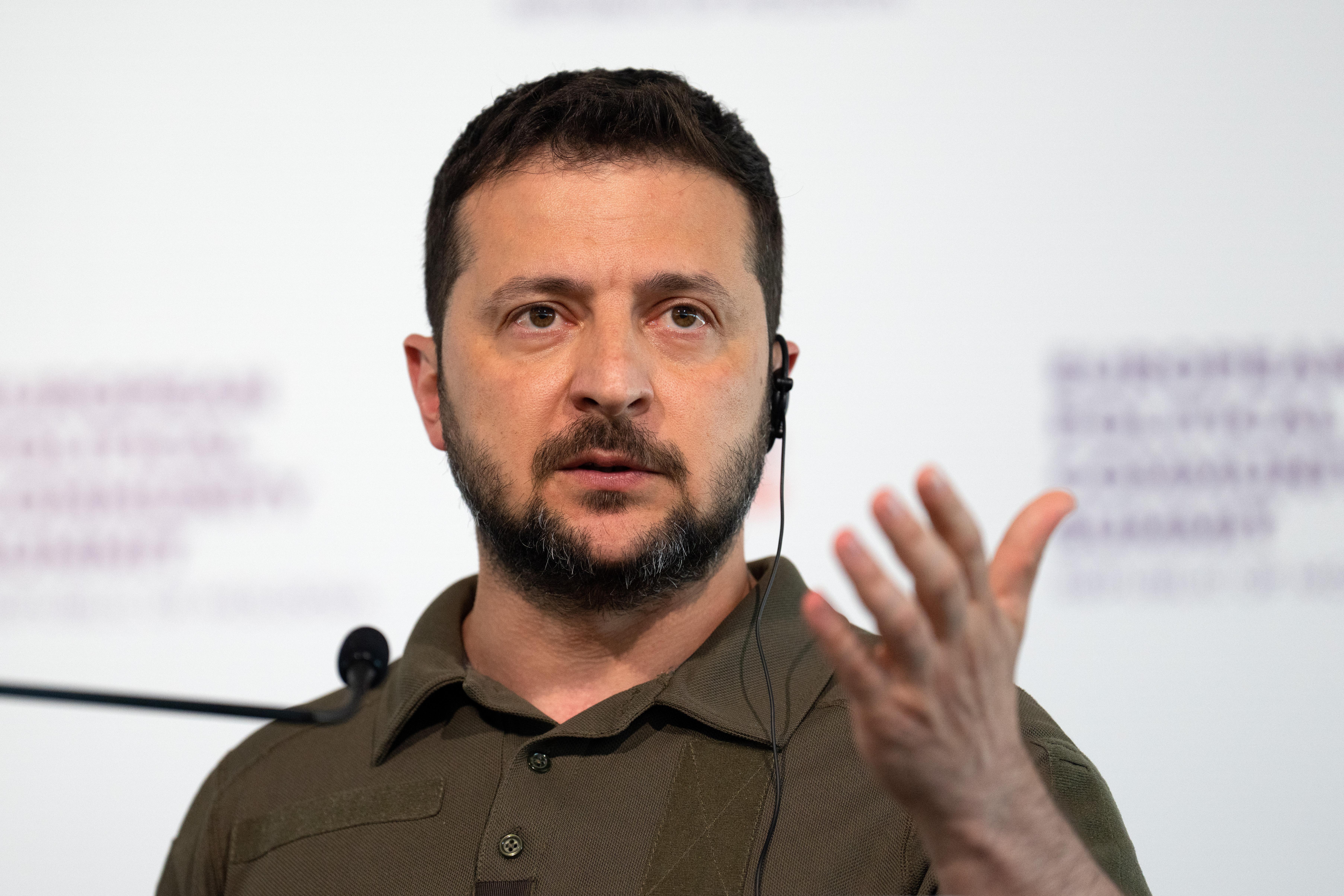 Volodymyr Zelensky, wearing a brown collared shirt, speaks before a microphone.