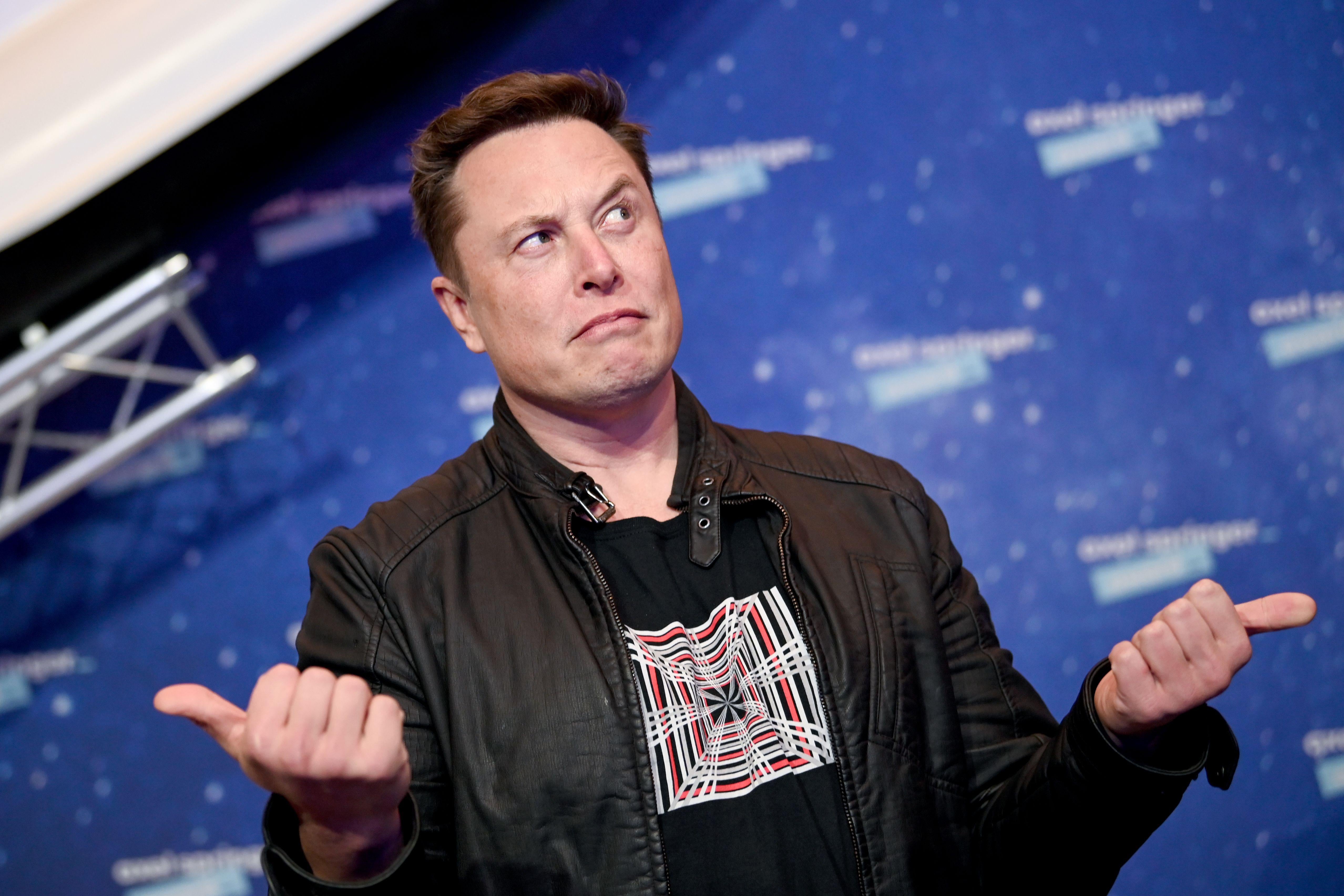 Elon Musk stands on a red carpet with a quizzical expression, making fists with both hands with the thumbs sticking out sideways