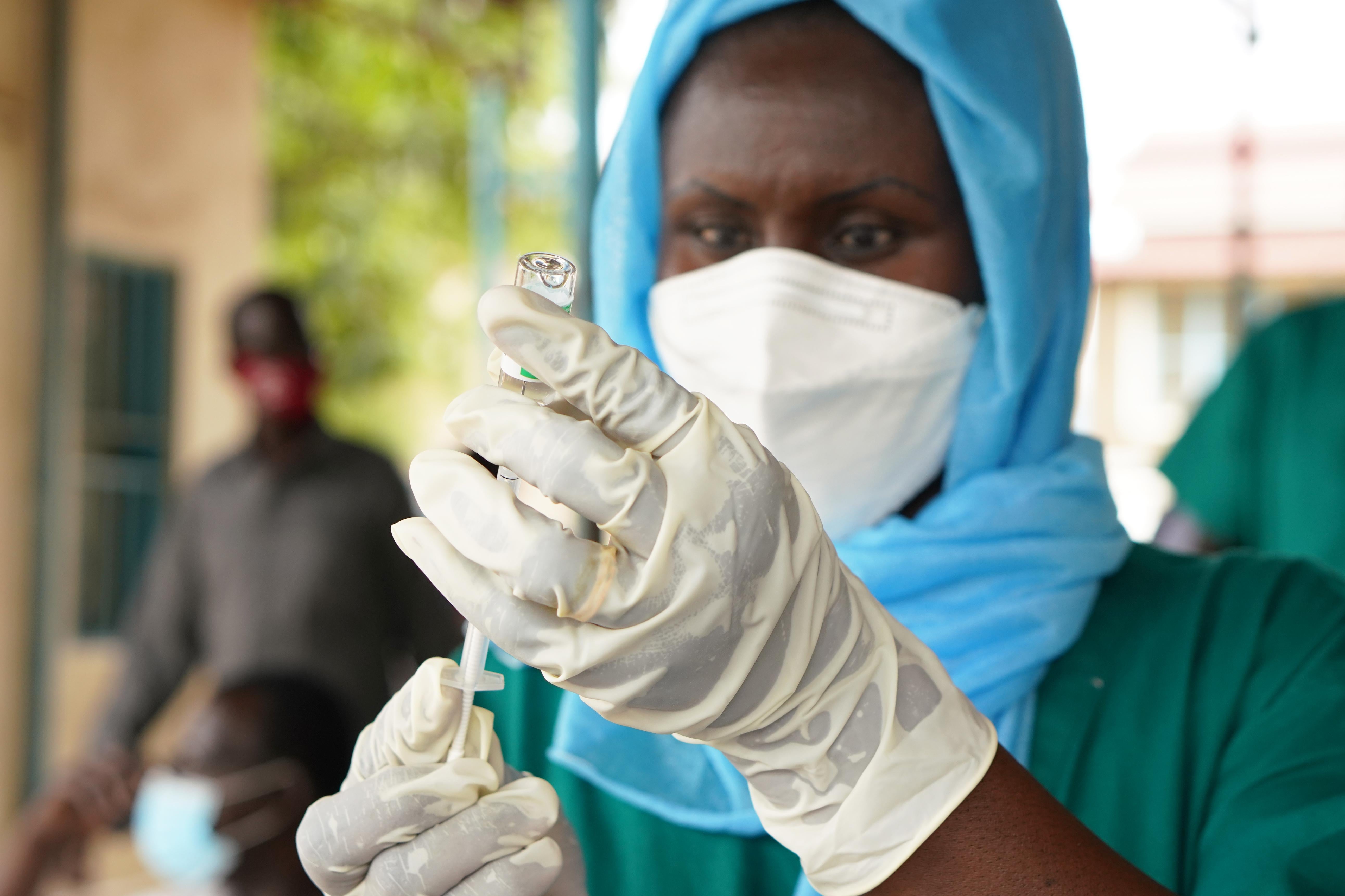 A person in a headscarf, face mask, and medical gloves draws vaccine from a vial into a syringe.