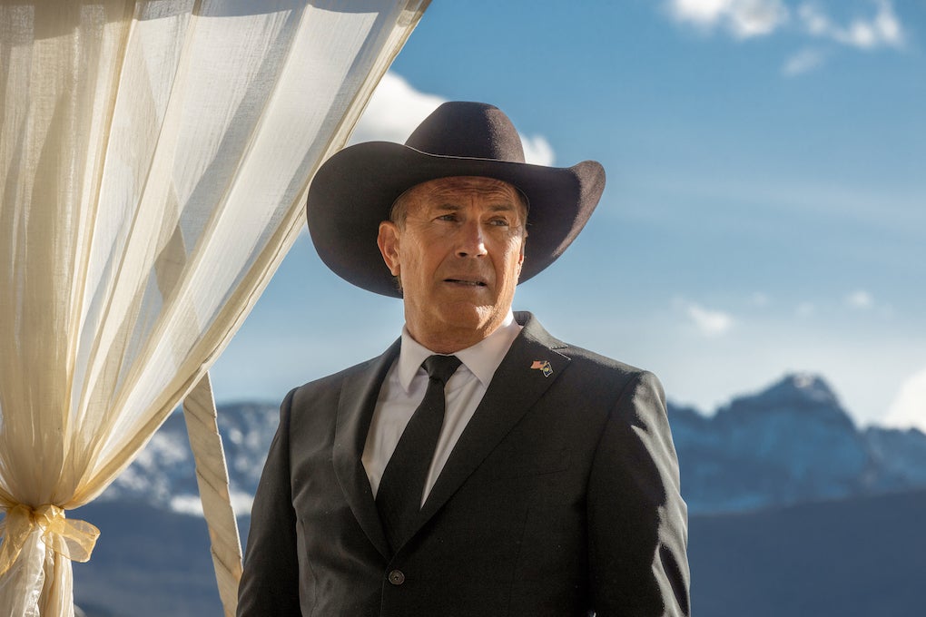 A man in a cowboy hat stands in front of a majestic mountain range.