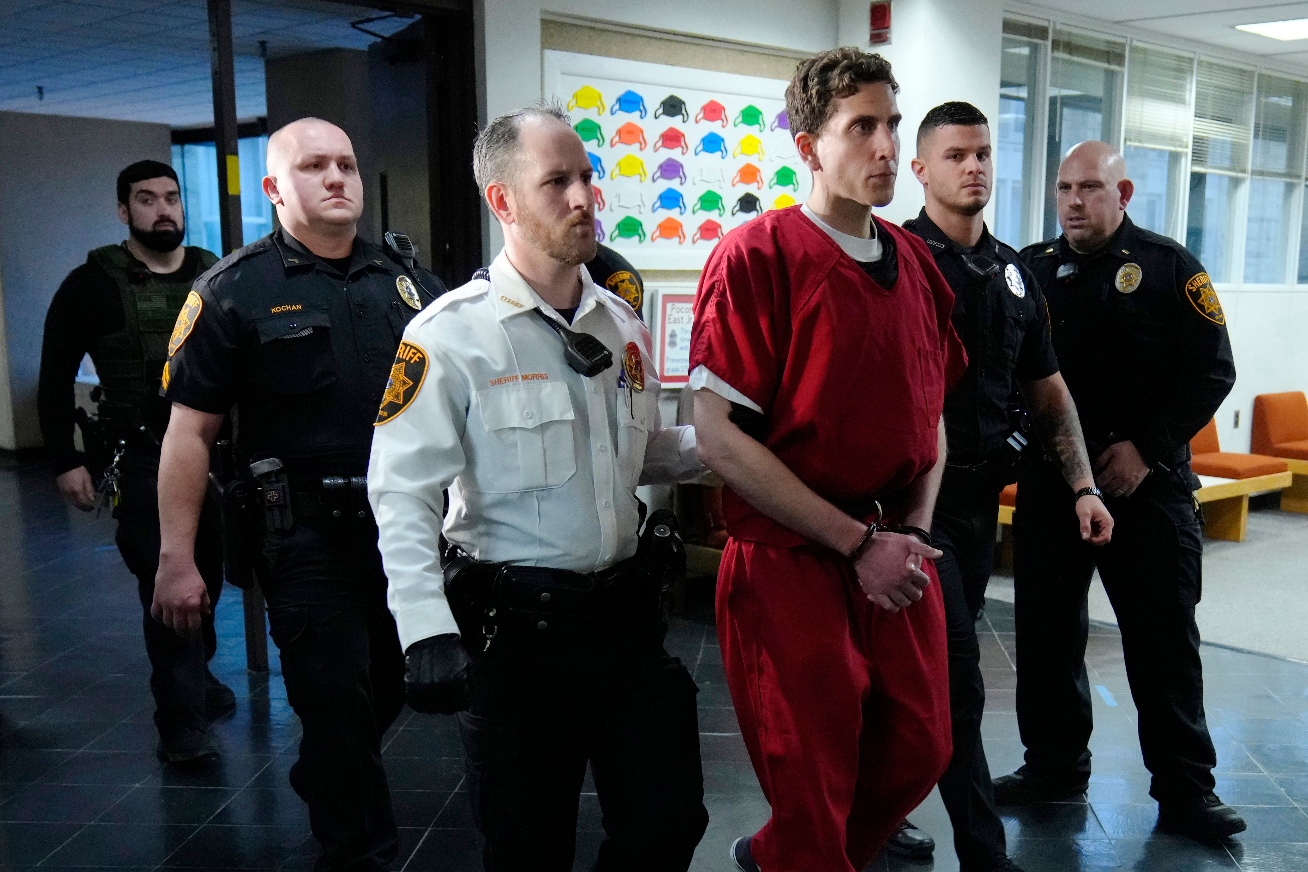 Kohberger, wearing an orange jumpsuit and handcuffs, is escorted down a hallway and flanked by several members of law enforcement.