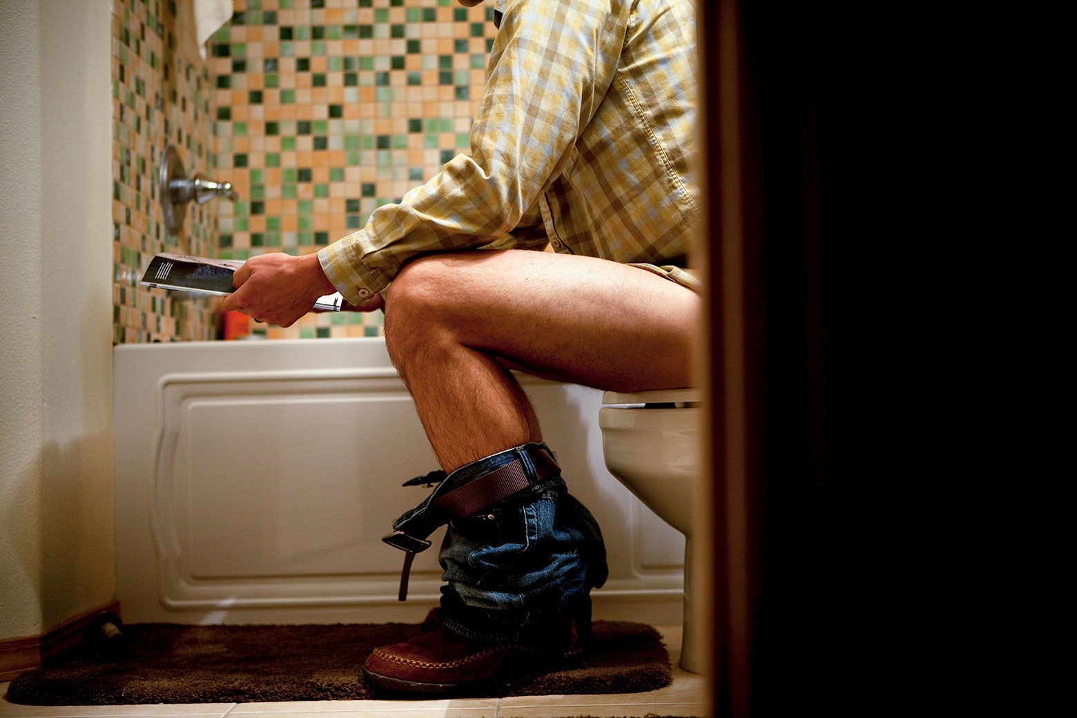 A man is seen from the side in his bathroom, pants down, sitting on a toilet, reading a magazine