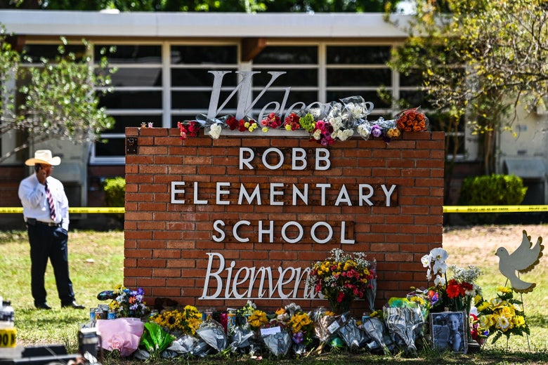 A sign that says "Welcome Robb Elementary School Bienvenidos" is covered with flowers; a man in a cowboy hat stands in the distance.